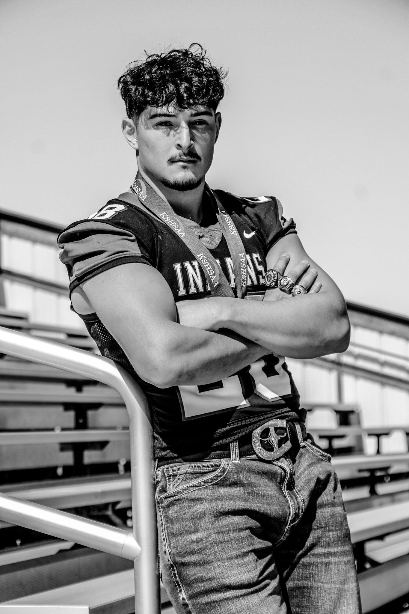 High school football player in uniform posing against stadium bleachers in black and white.