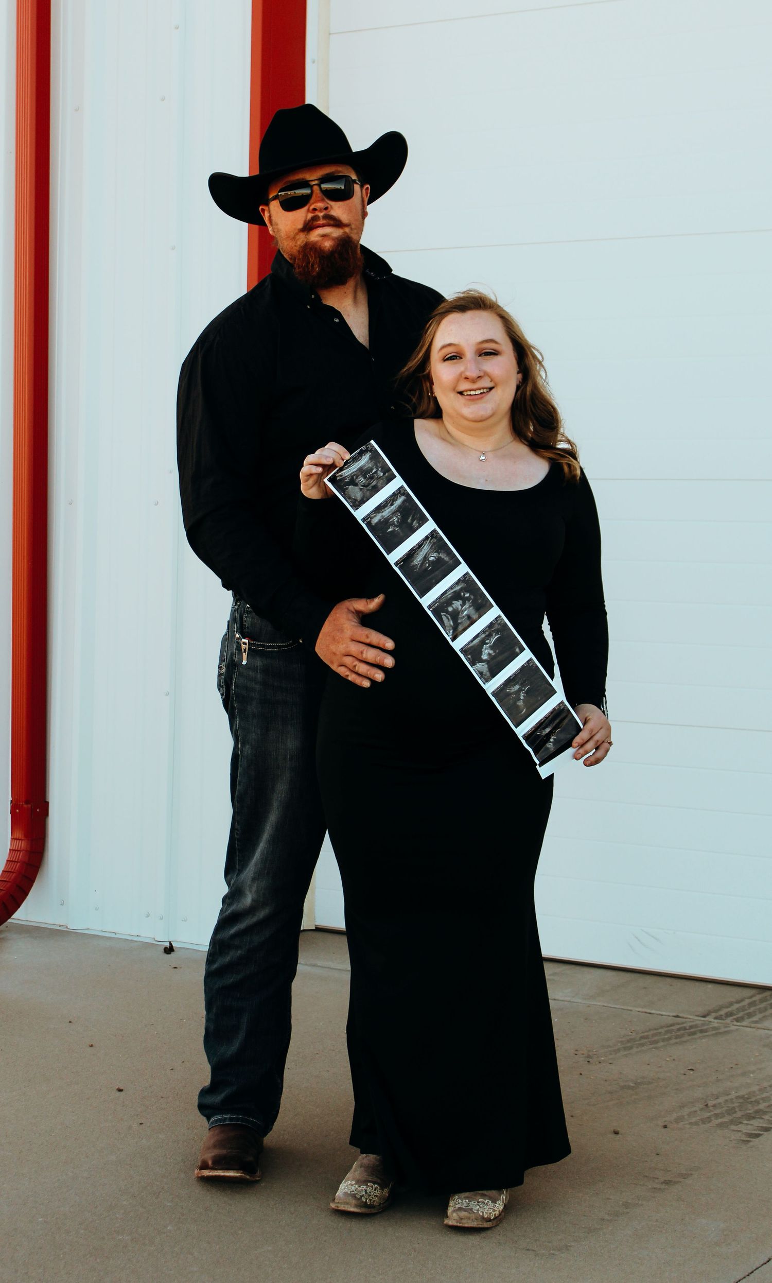 A couple poses outdoors with ultrasound photos while holding a black and white strip of sonogram images.