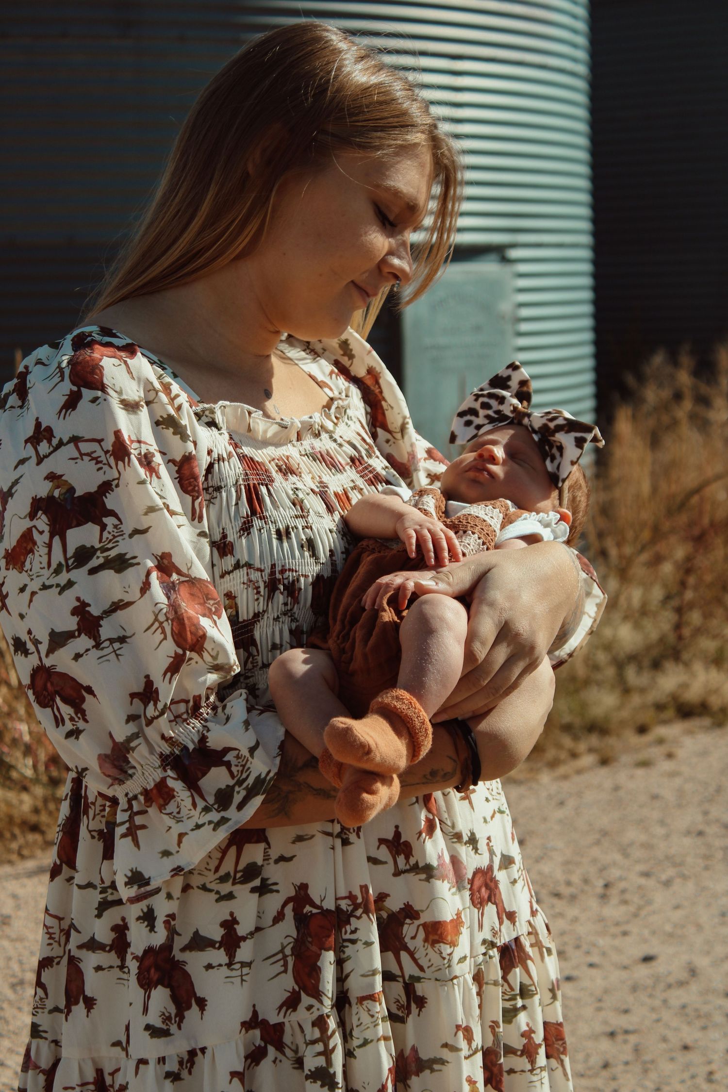 Woman in floral print dress holds baby outdoors in warm sunlight against metal siding wall.