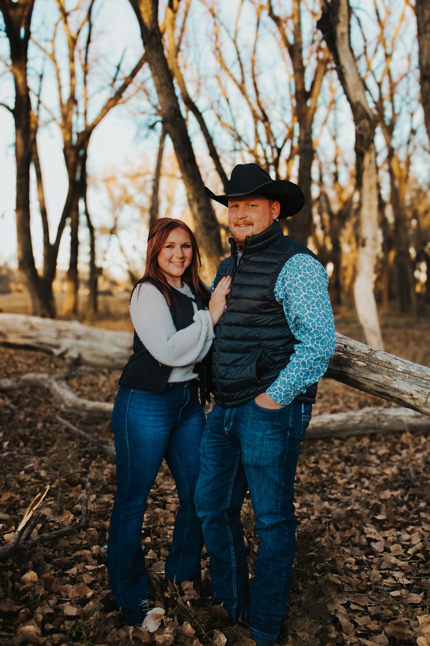 A couple in western attire posing together in a wooded setting during golden hour.