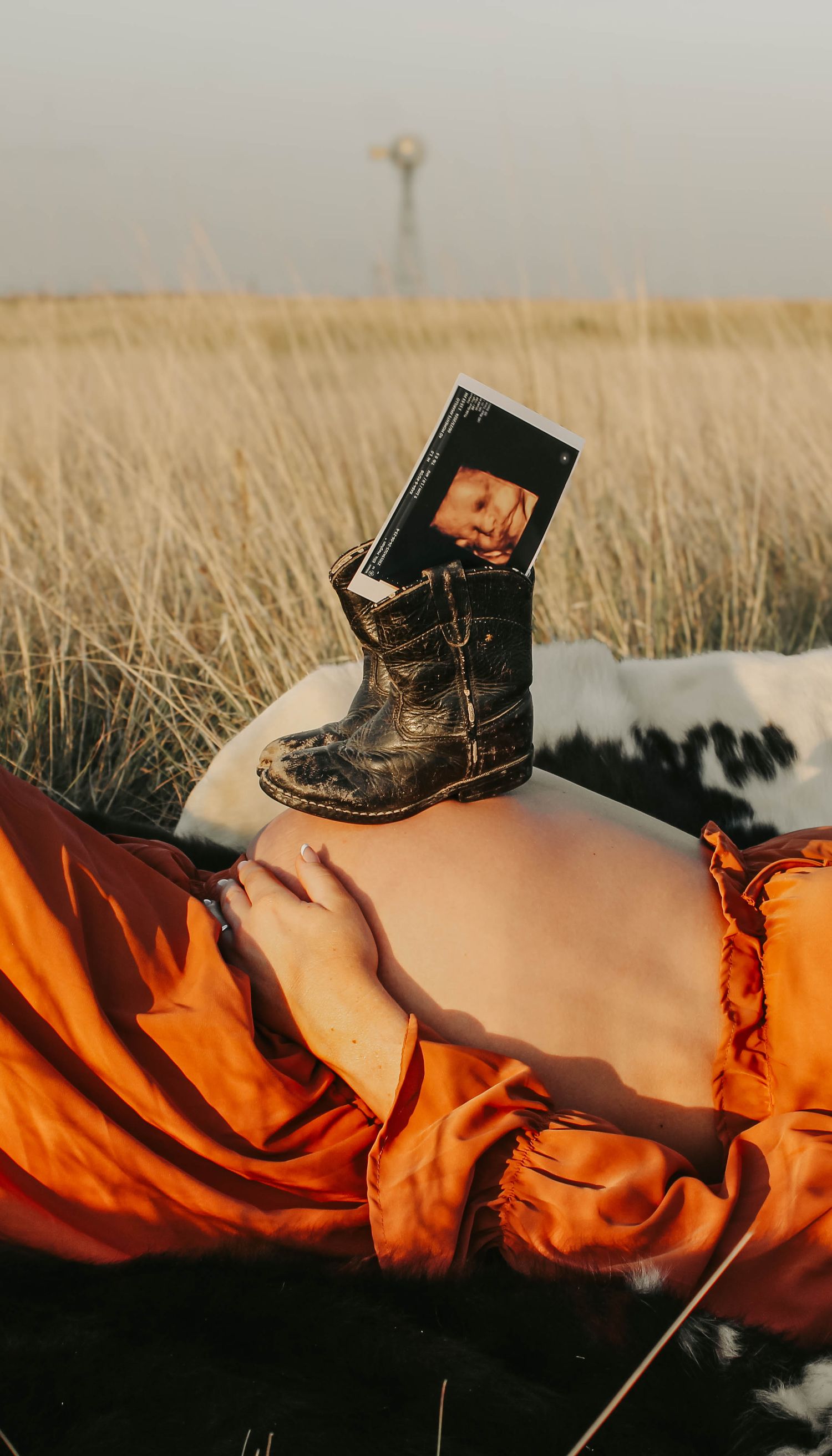 Artistic maternity photo featuring a black boot holding a photo in tall grass at sunset.