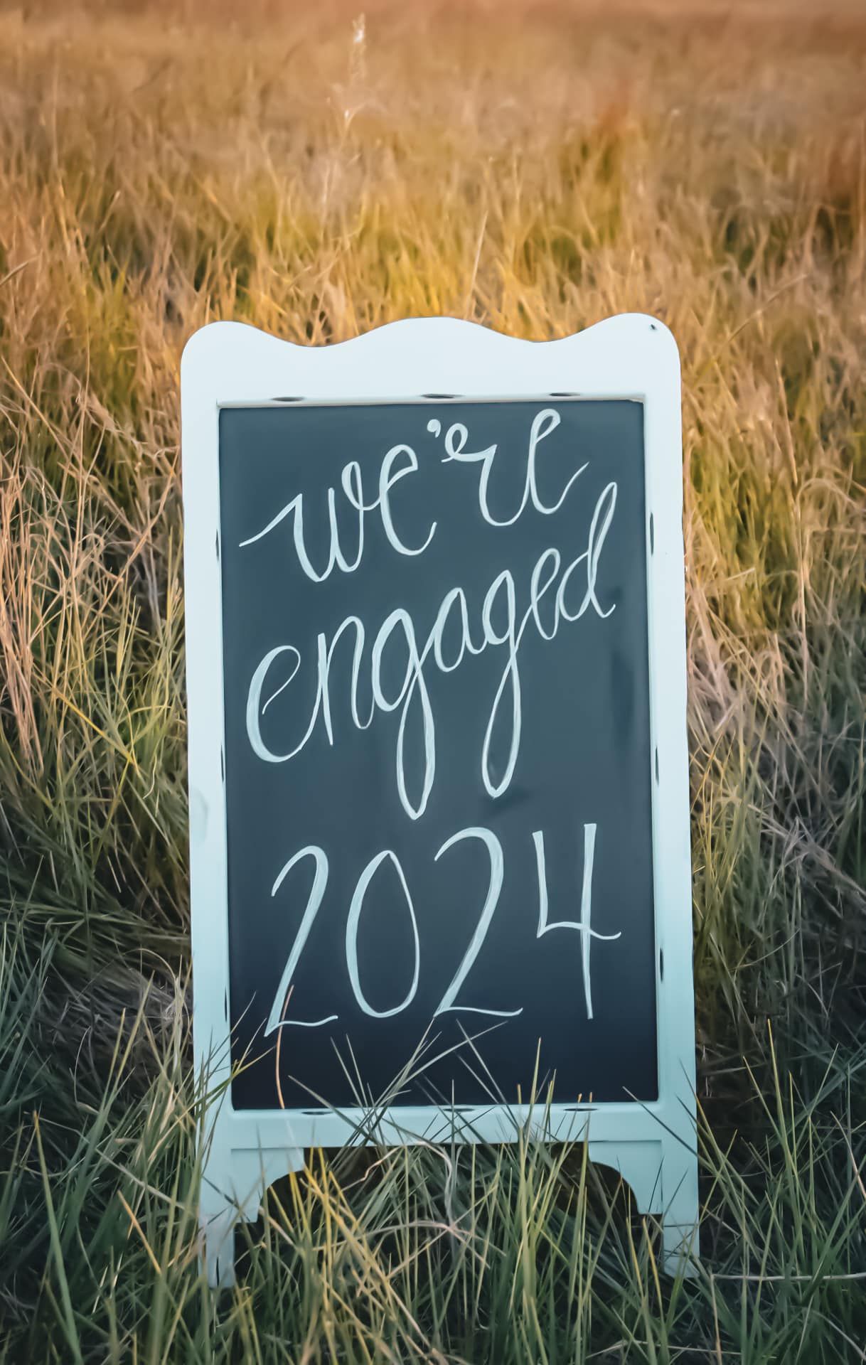 A white chalkboard sign in a wheat field announces 'We're engaged 2024'.