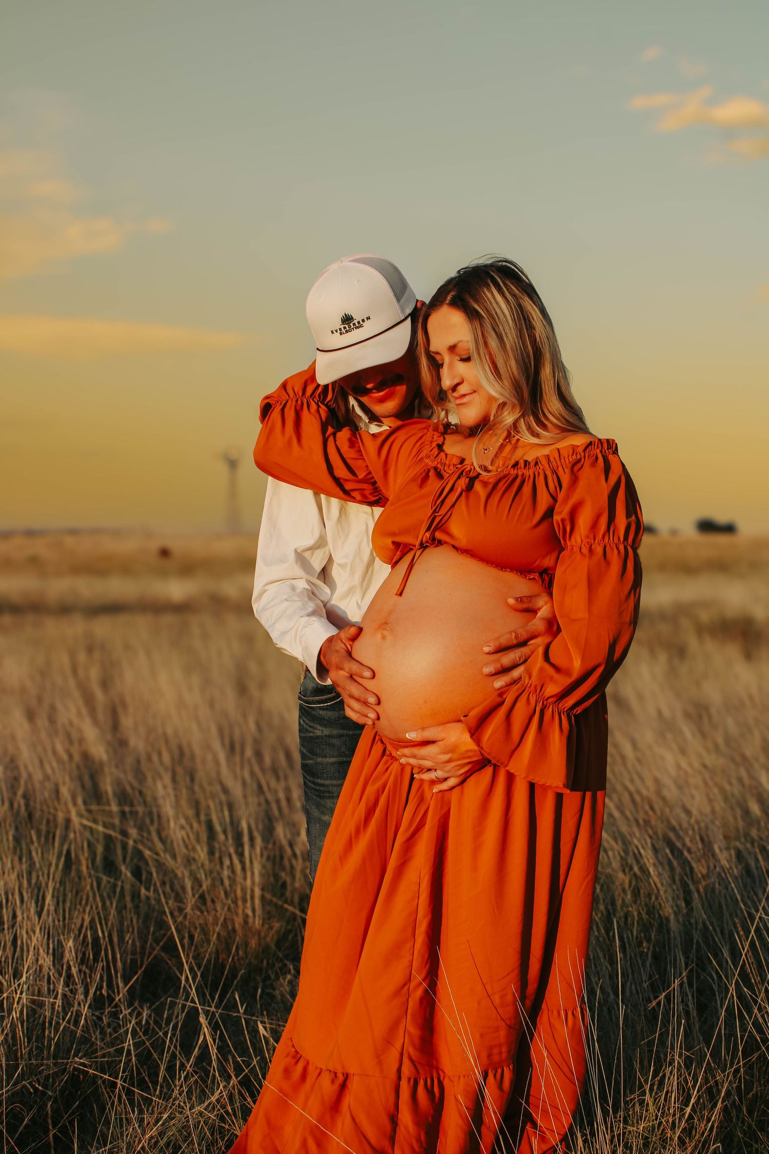 A couple embraces in a wheat field at sunset, showing off a baby bump in an orange dress.