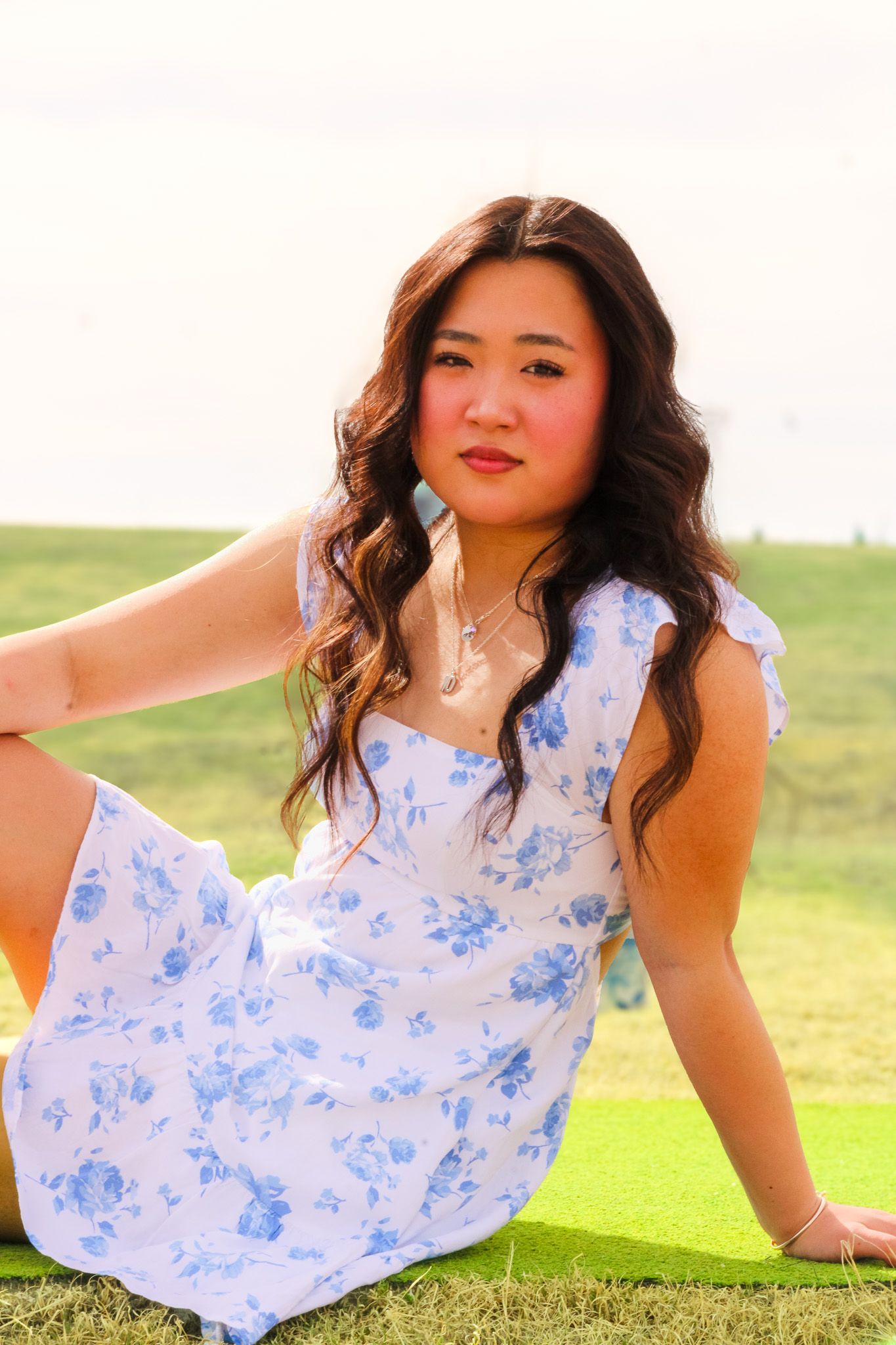A person in a white and blue floral dress poses gracefully on grass during a sunny outdoor photoshoot.
