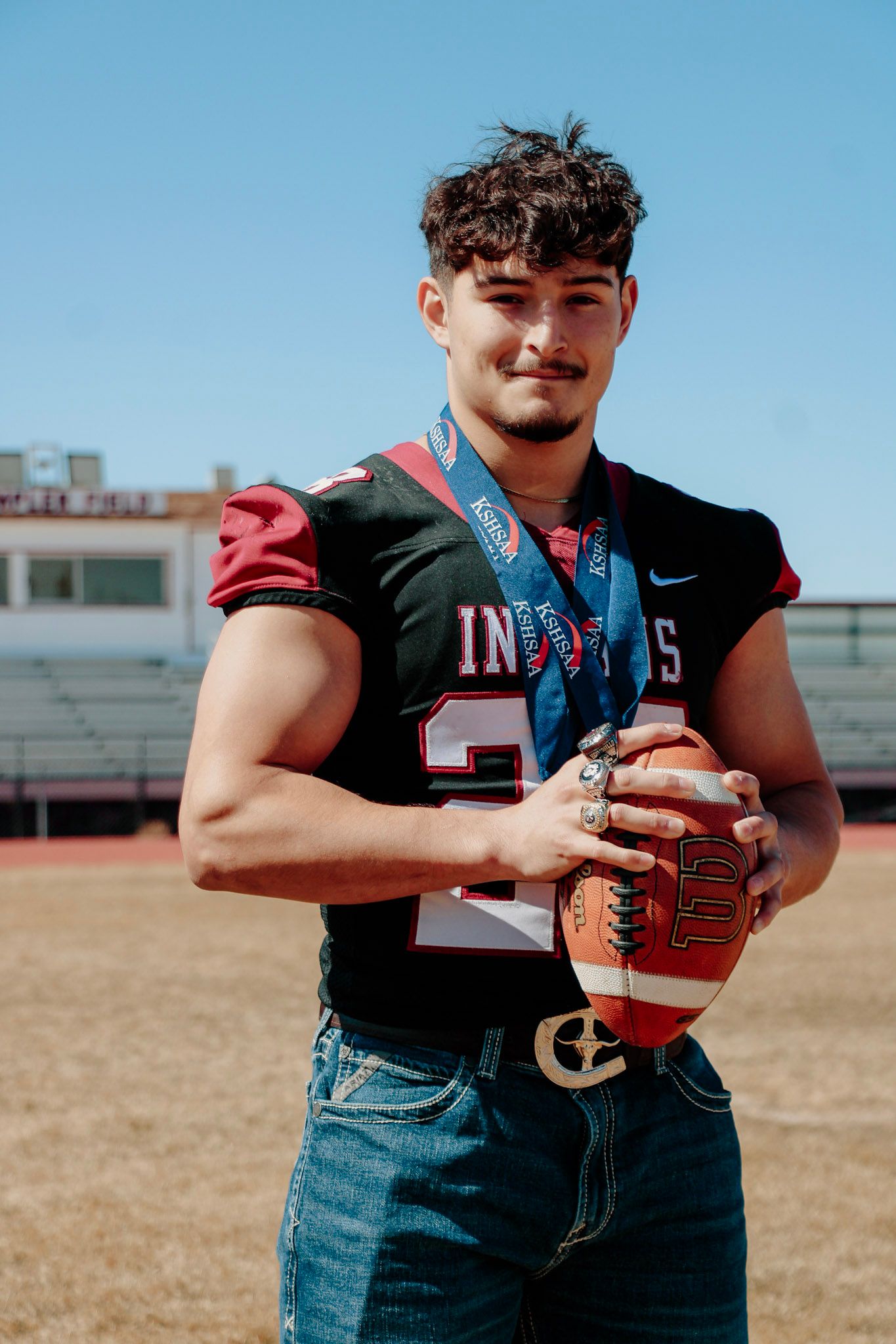 A football player in a black jersey and jeans holds a football against a sunny outdoor field background.