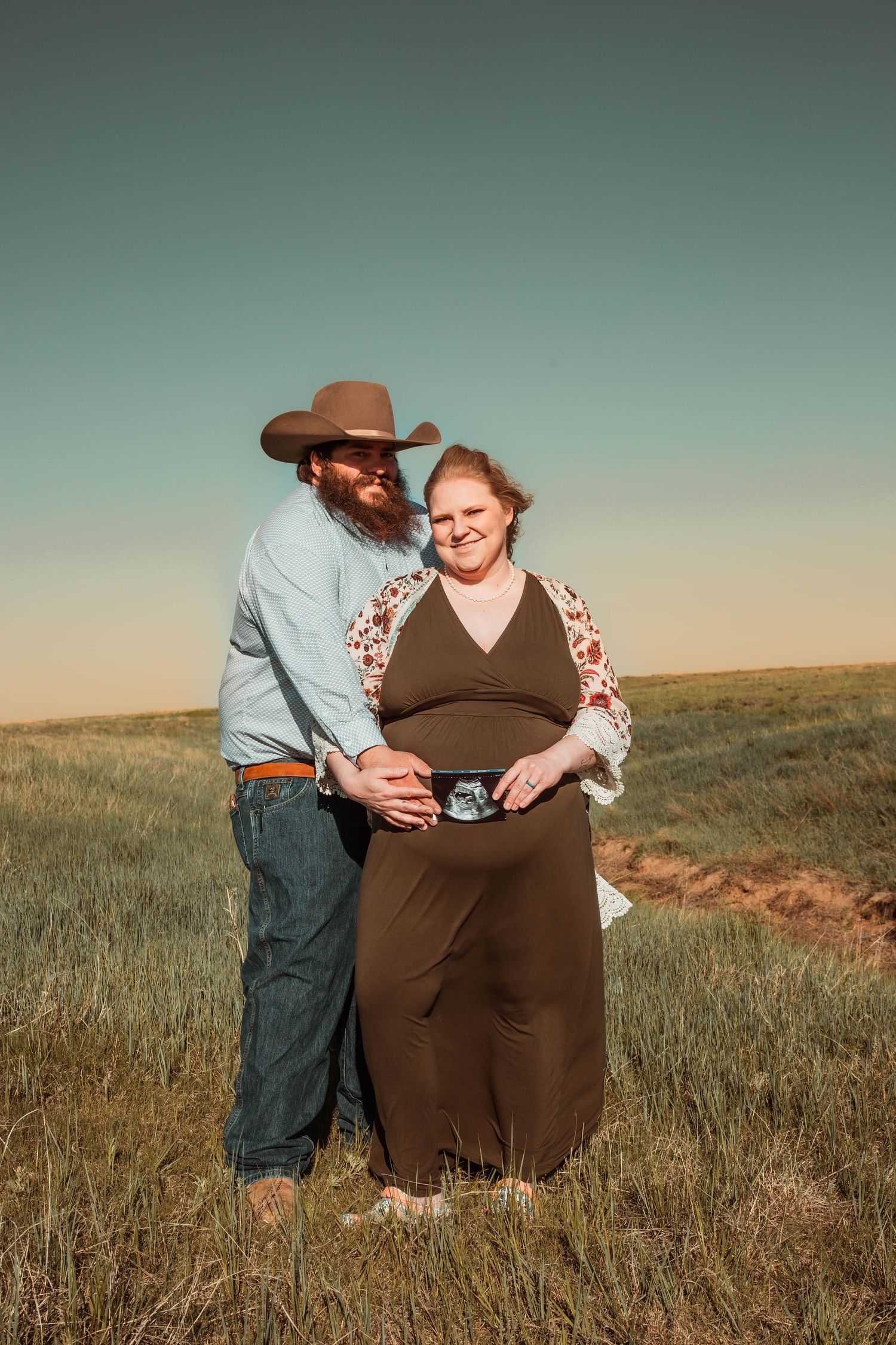 A couple stands together in a grassy field at sunset as they hold a sonogram photo in front of a brown dress.