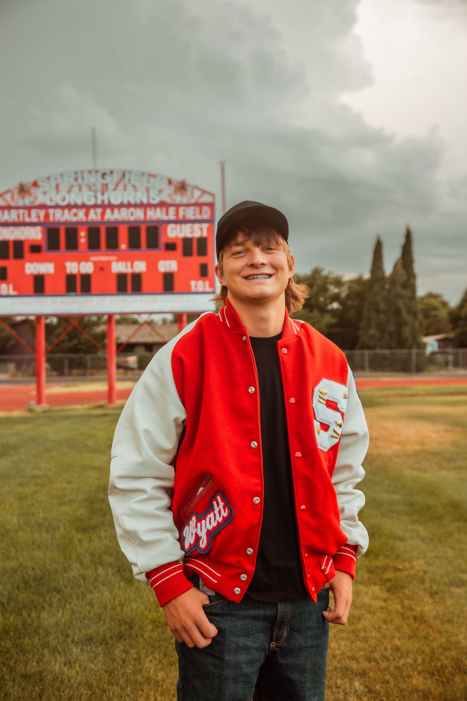 Red and white varsity letterman jacket shown against backdrop of high school baseball field and stadium.