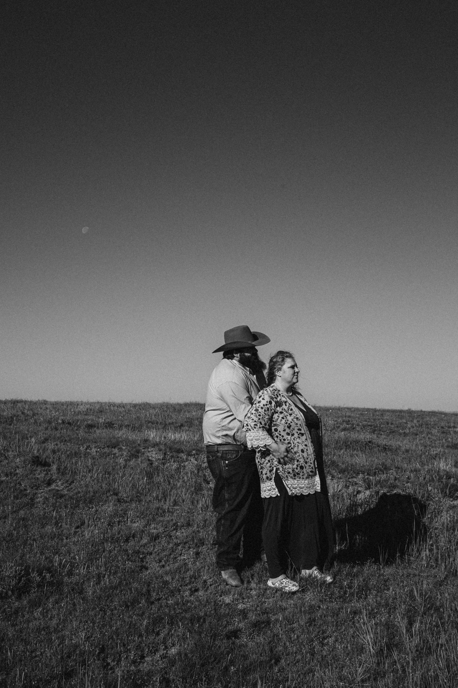 A black and white photo of a couple embracing on a grassy hillside under an open sky.