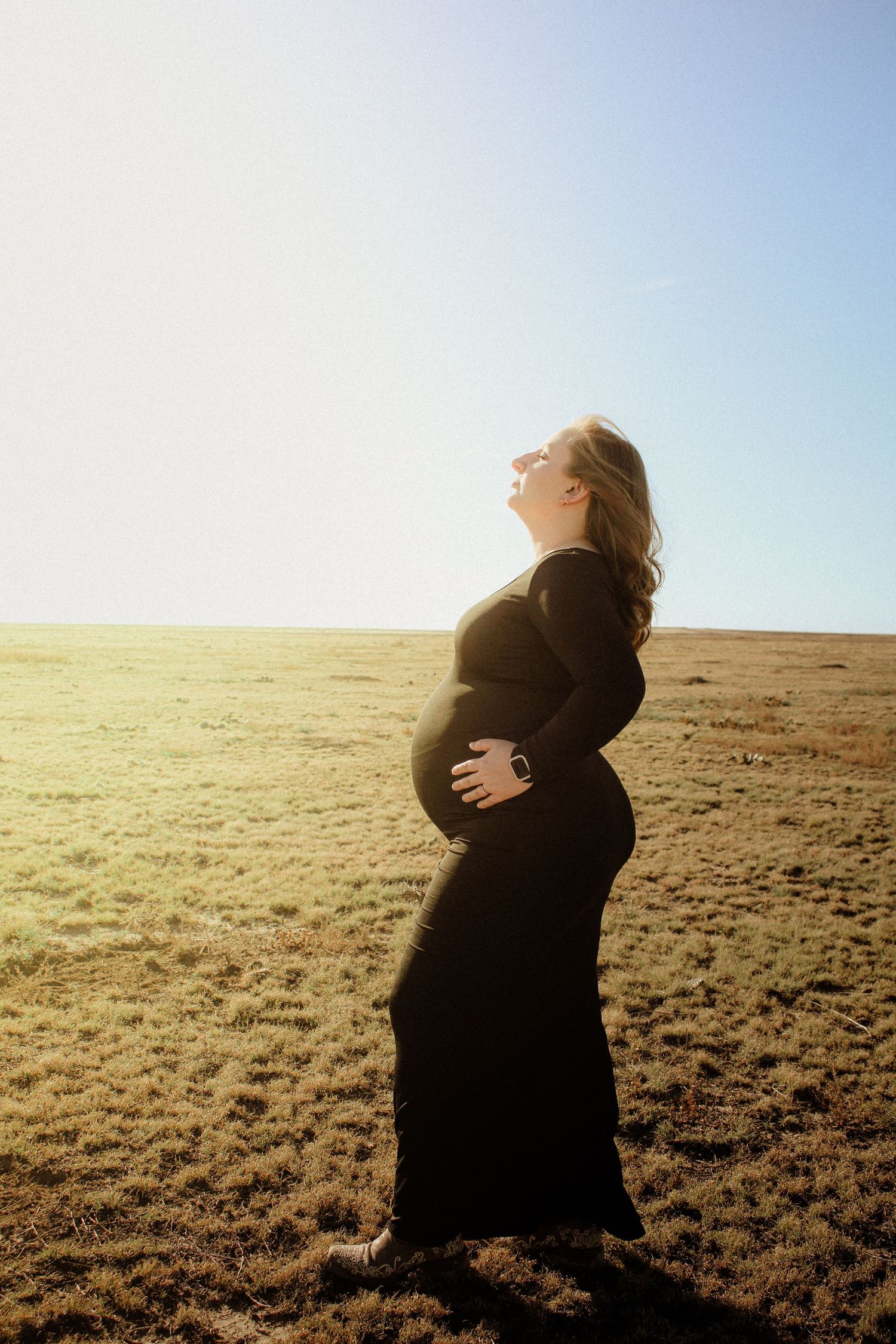 Silhouetted pregnant woman in long black dress stands in sunlit field during golden hour.