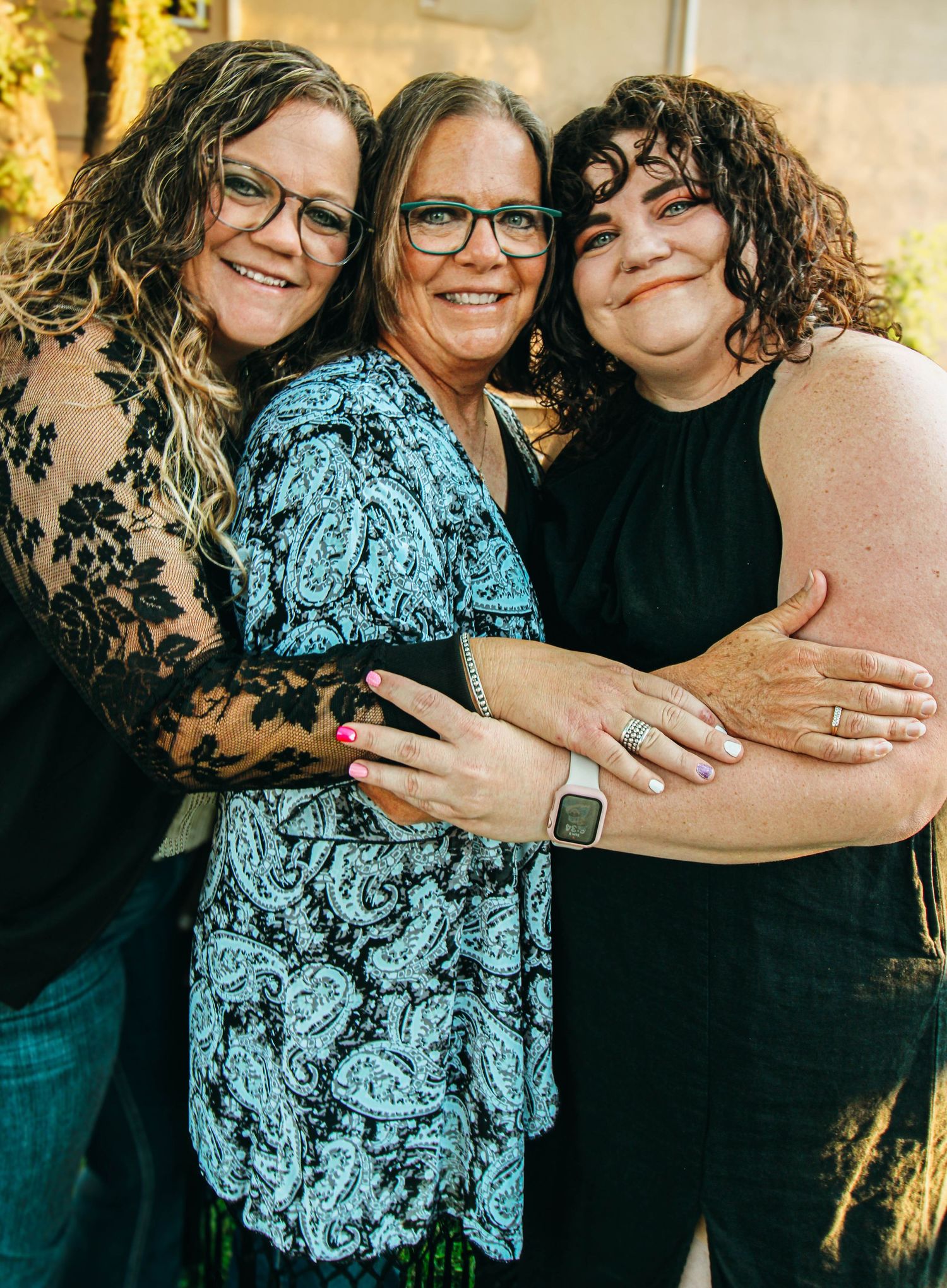 Three women embrace while smiling warmly in outdoor evening light.