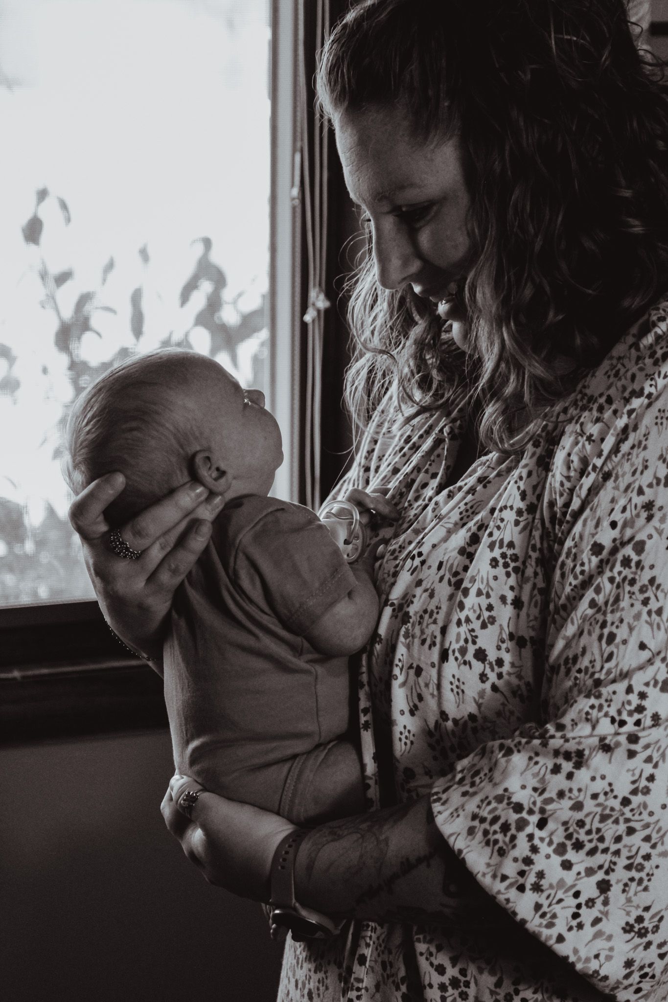 A tender black and white moment shows an adult cradling a baby near a window with patterned shadows on the wall.