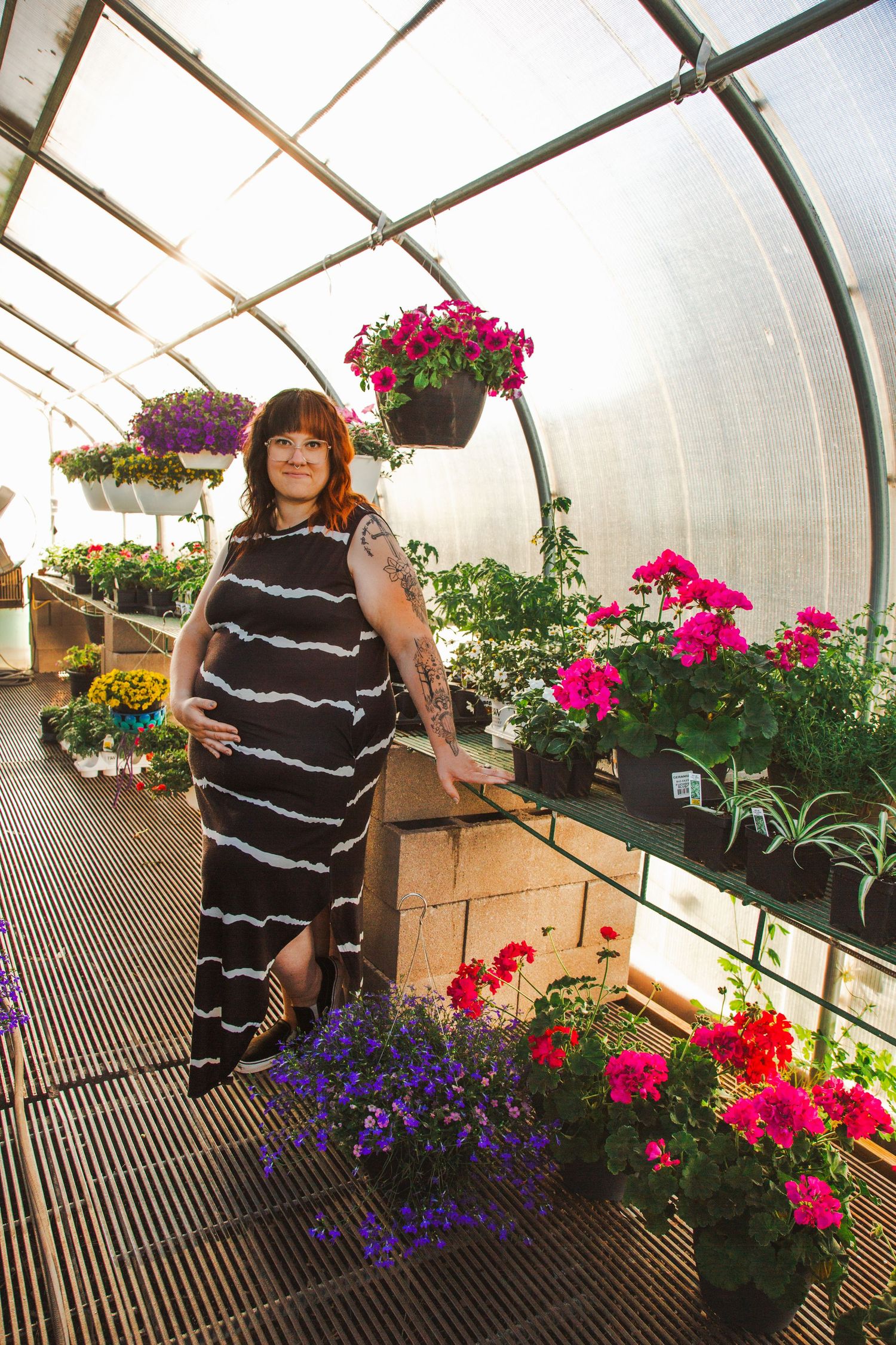 A person in a striped maxi dress stands among colorful hanging flower baskets and potted plants in a sunny greenhouse.