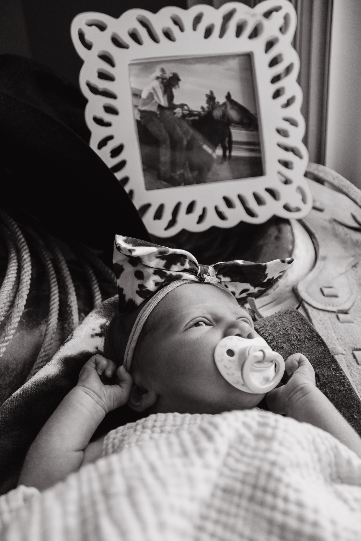 Black and white portrait of sleeping baby with pacifier under framed photograph on wall.