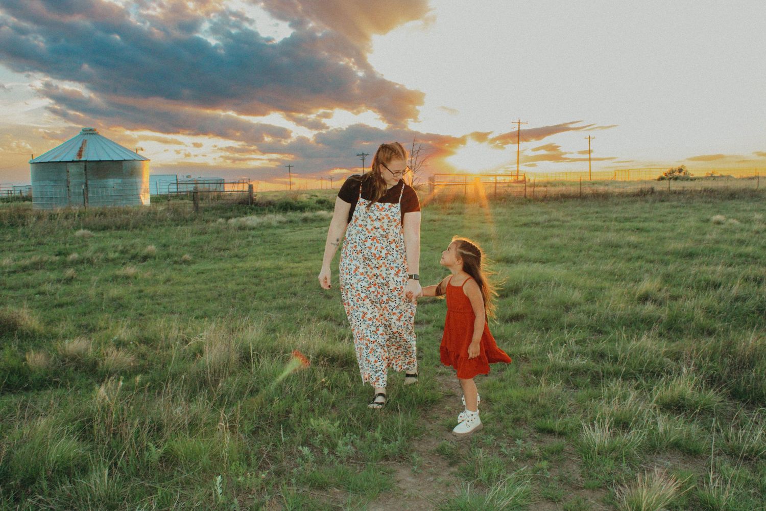 A parent and child walk through a rural field at sunset with farm buildings in the background.
