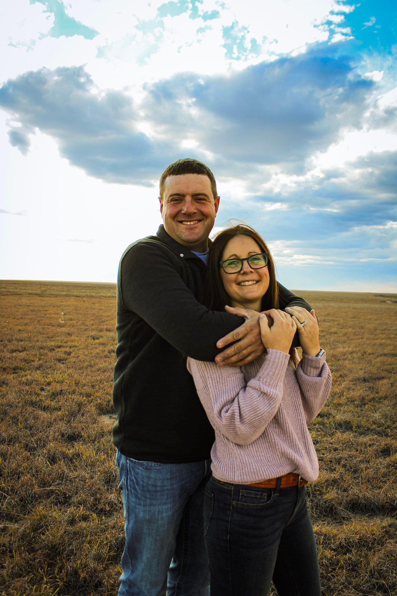 Happy couple embracing in casual sweaters against vast prairie landscape with dramatic sky.