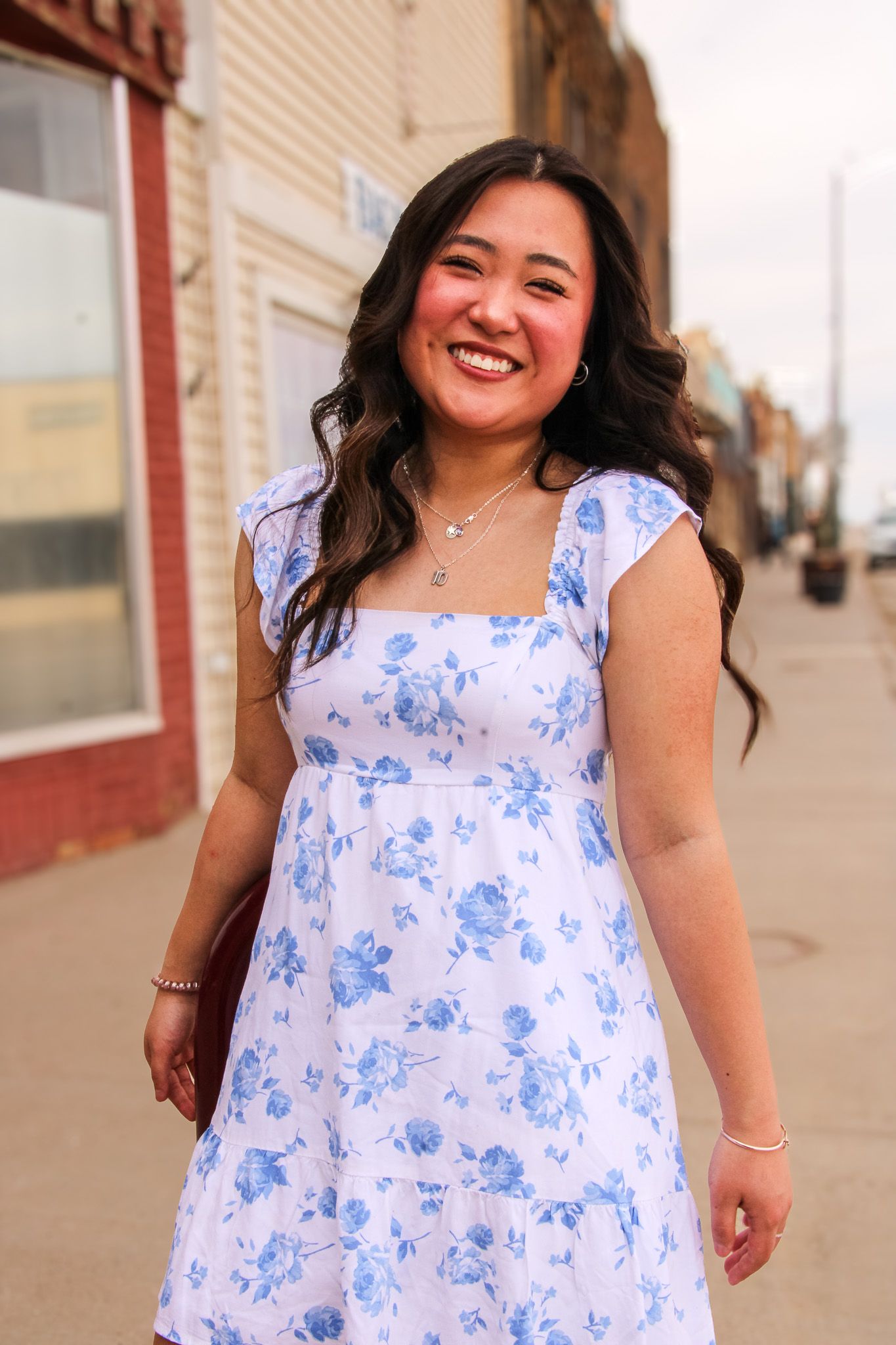 A person wearing a white and blue floral print dress stands smiling on a sidewalk near brick buildings.