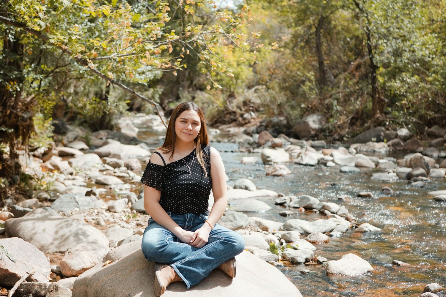 Young woman sitting cross-legged on a rock by a peaceful forest stream, wearing a black top and jeans.