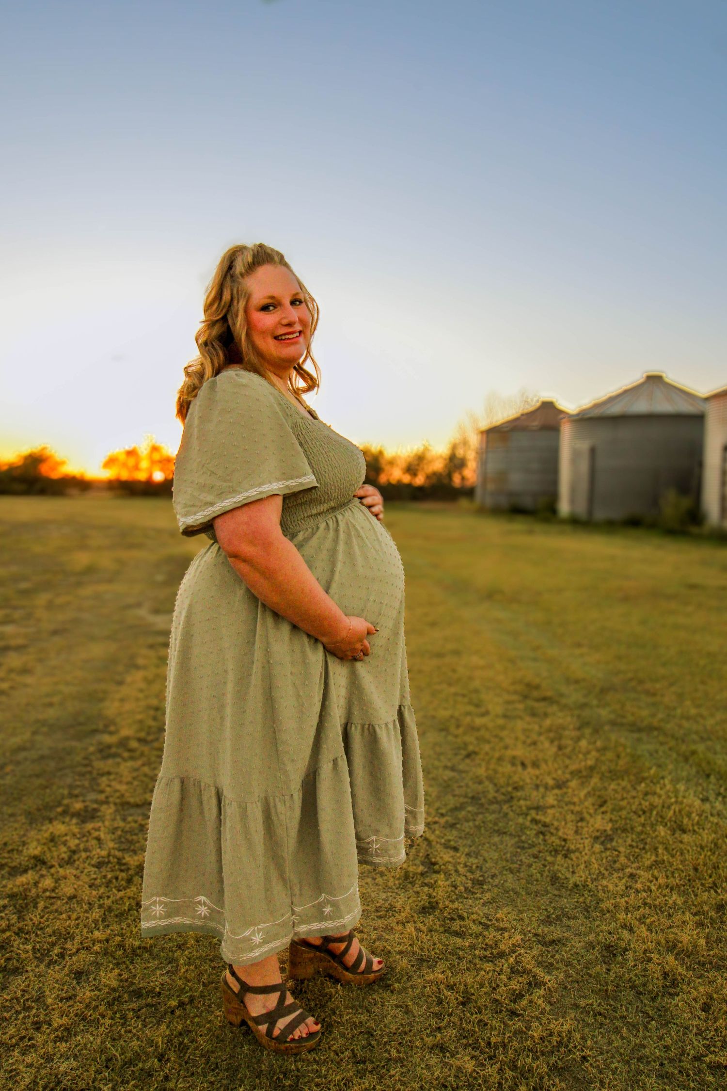 An expectant person in a sage green dress poses in a field at sunset with grain bins in the background.