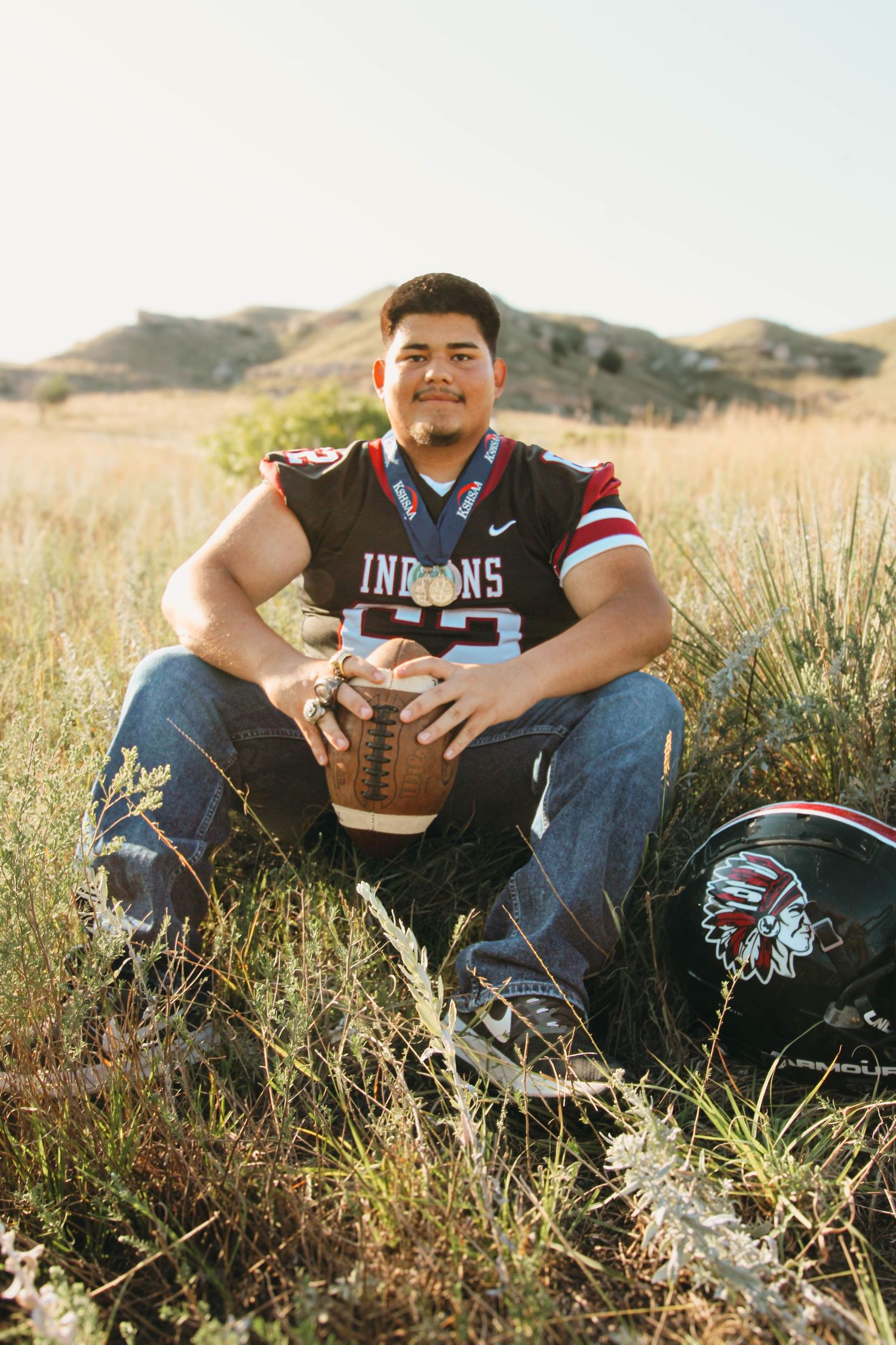 Football player sits in tall grass with a football and sports jersey during an outdoor photo session.