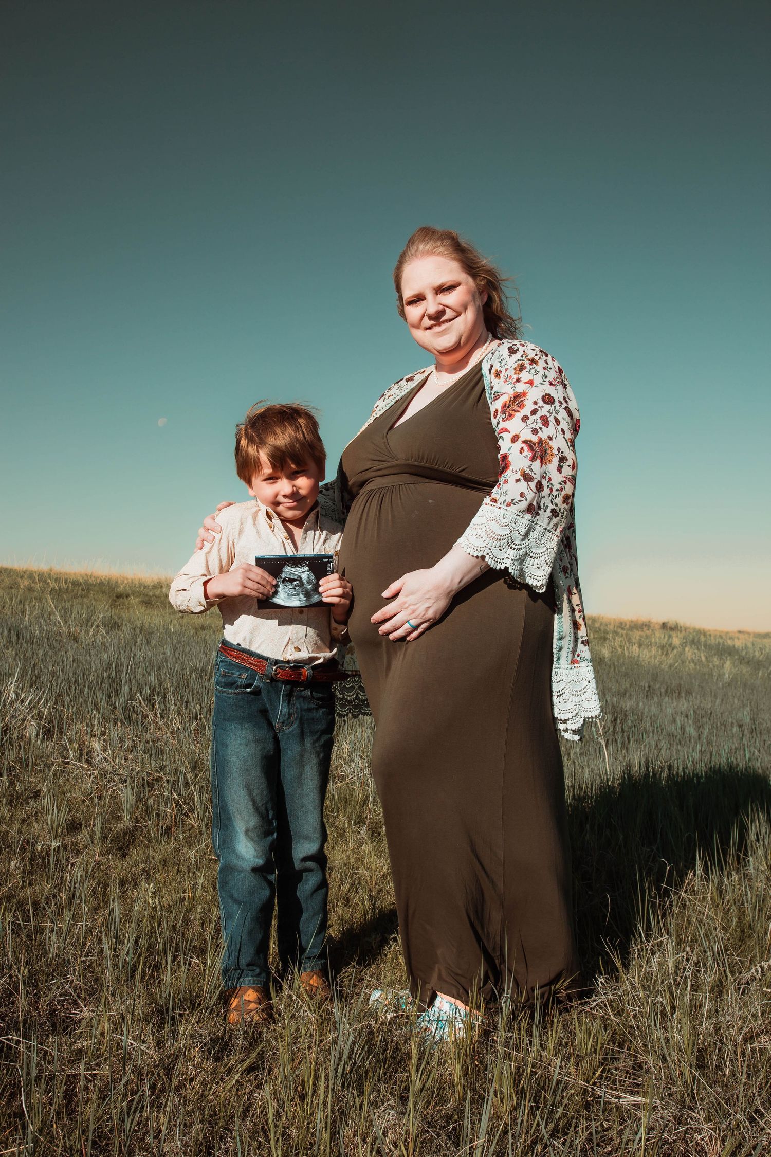 A pregnant person in a brown dress stands in a grassy field with a child holding a camera under blue sky.