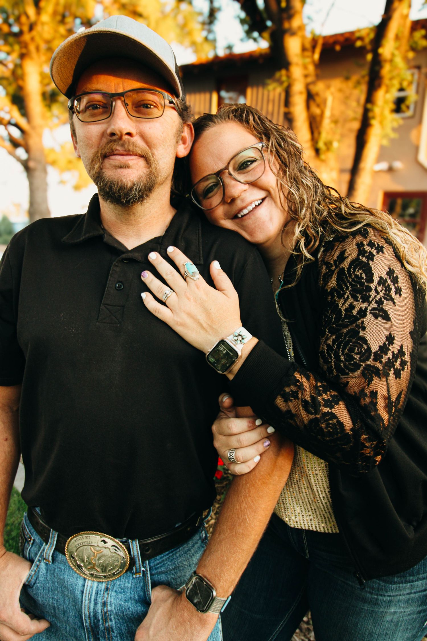Couple in casual attire embracing and smiling outdoors with fall foliage in background.