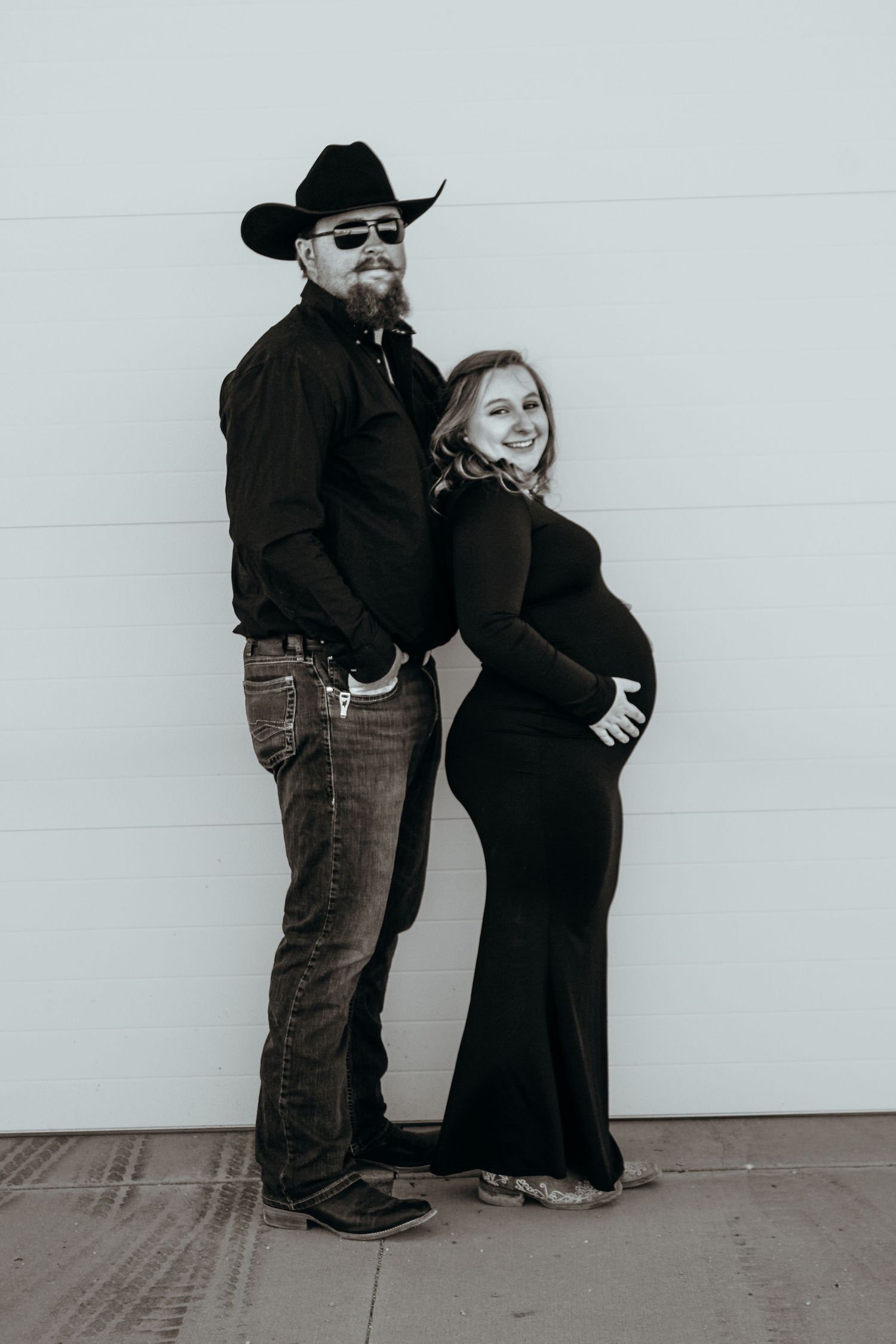 Black and white maternity photo showing a couple in western attire posing against a plain wall.