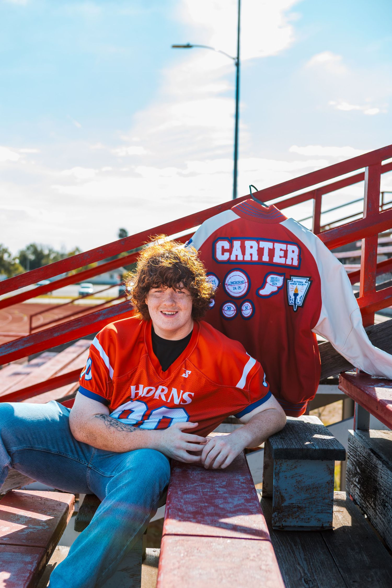 Teen boy in red Horns football jersey sits on bleachers next to a Carter banner on a sunny day.