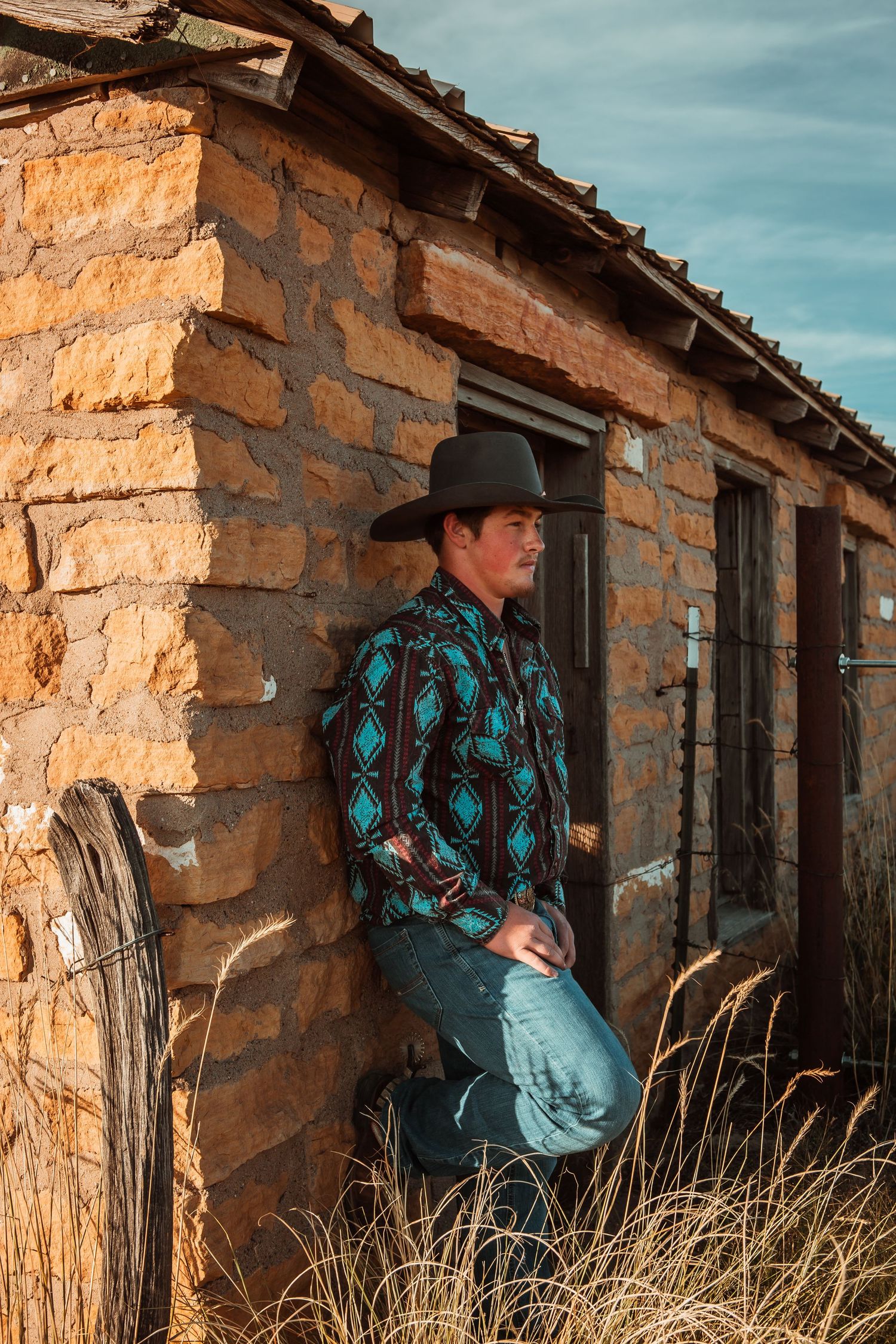 A cowboy wearing a turquoise western shirt and hat leans against a rustic stone building in warm sunset light.