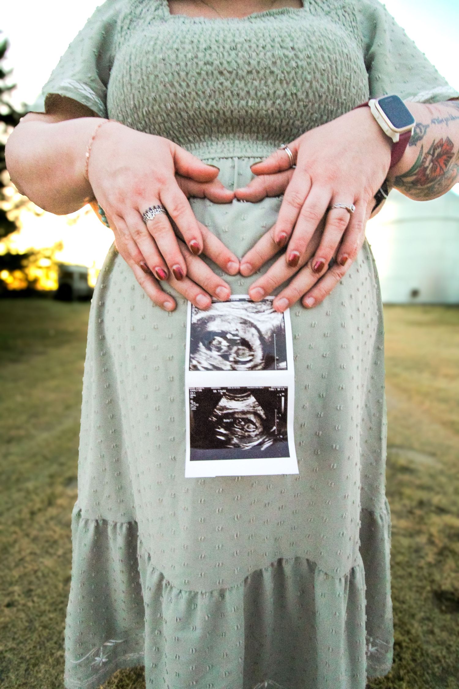 Someone in a sage green dress holds ultrasound photos while making a heart shape with their hands over their pregnant belly.