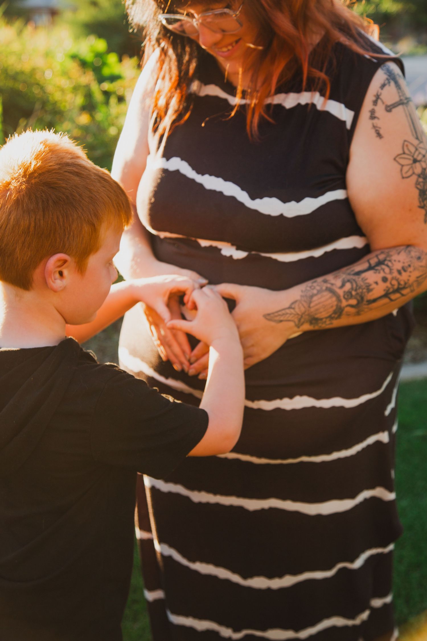 Child touches pregnant woman wearing black and white striped dress in warm sunlit outdoor setting.
