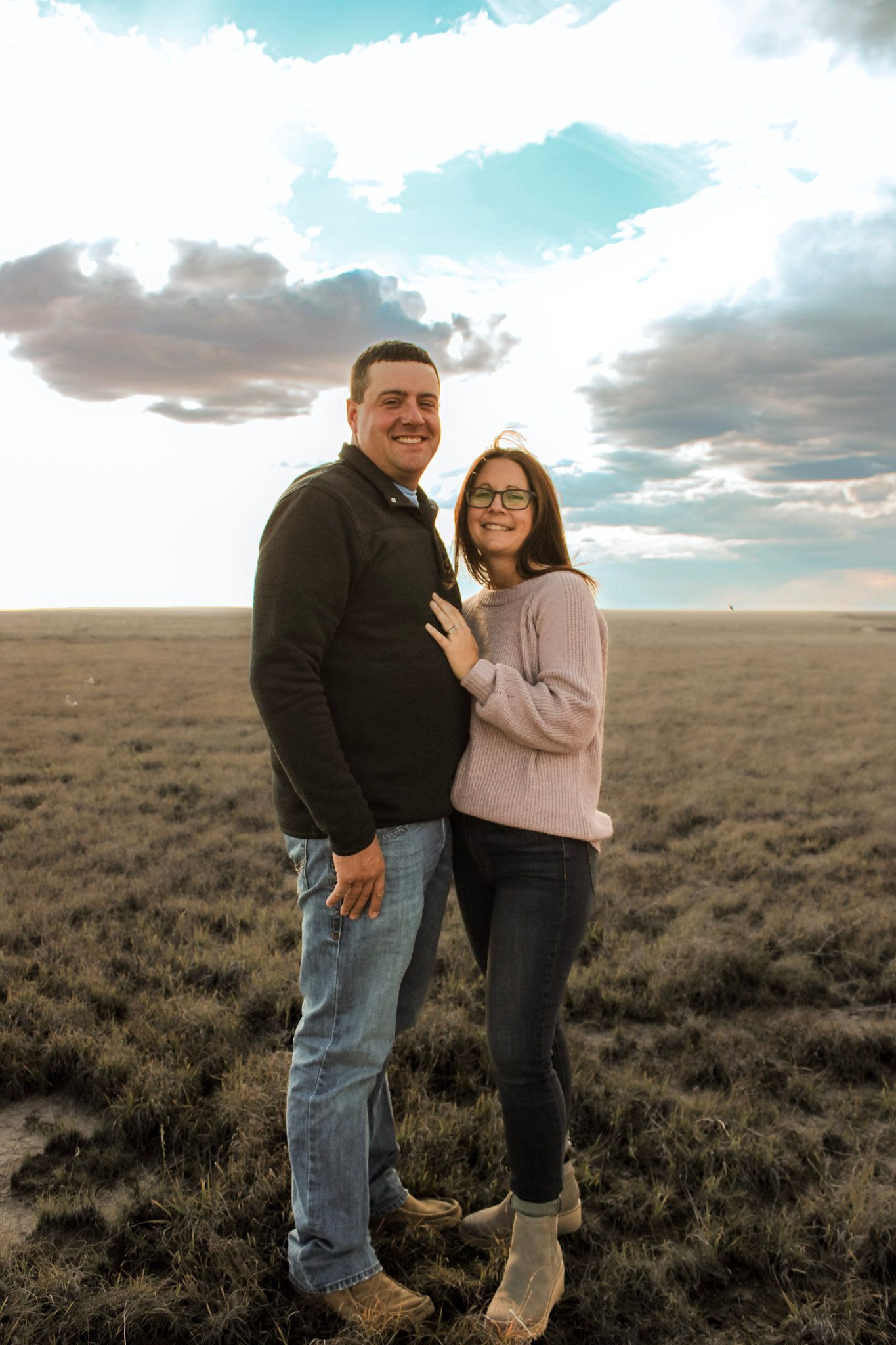 Couple embracing in an open field under dramatic cloudy sky at sunset.