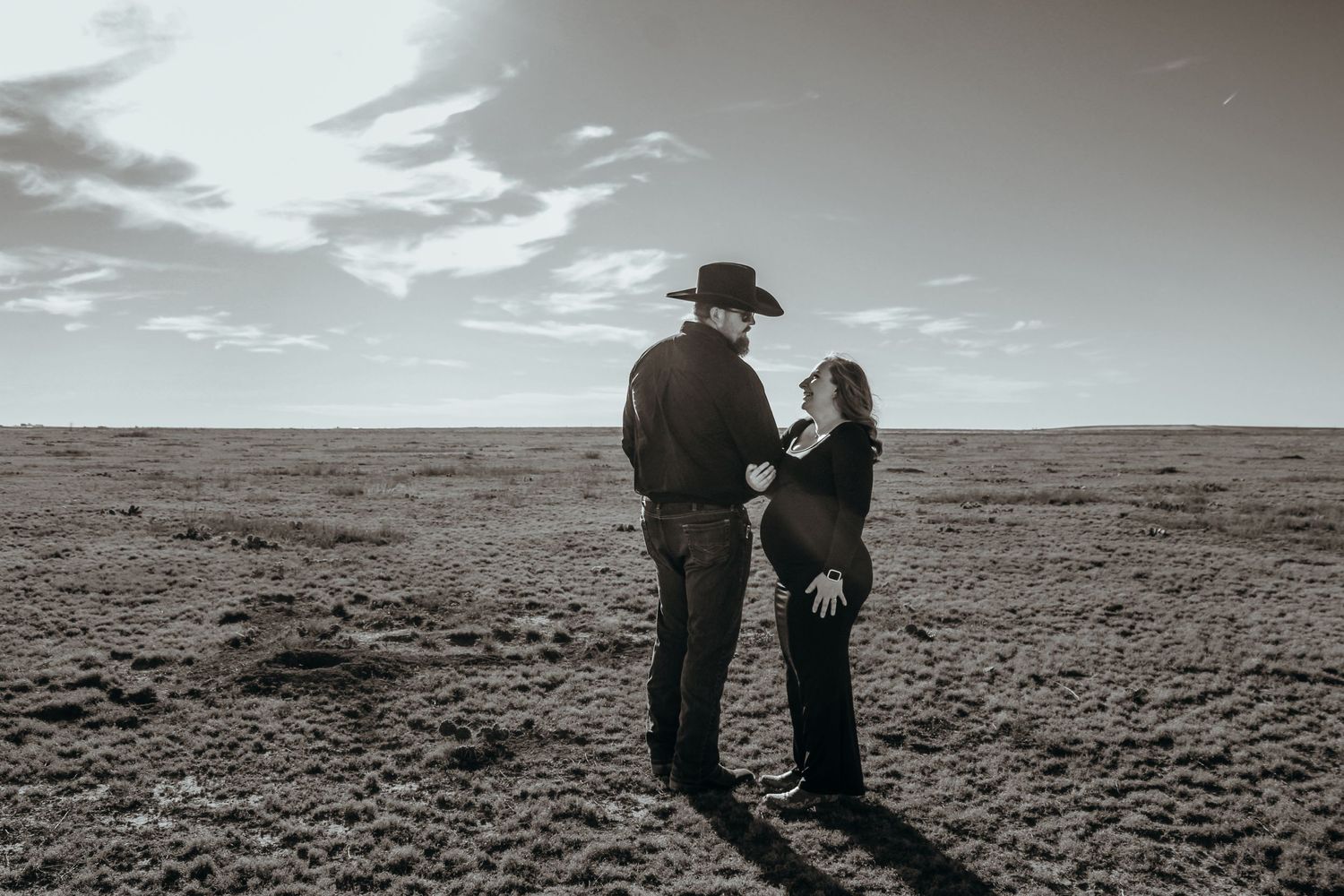 Two people stand in silhouette on desert terrain under cloudy skies in black and white photography.