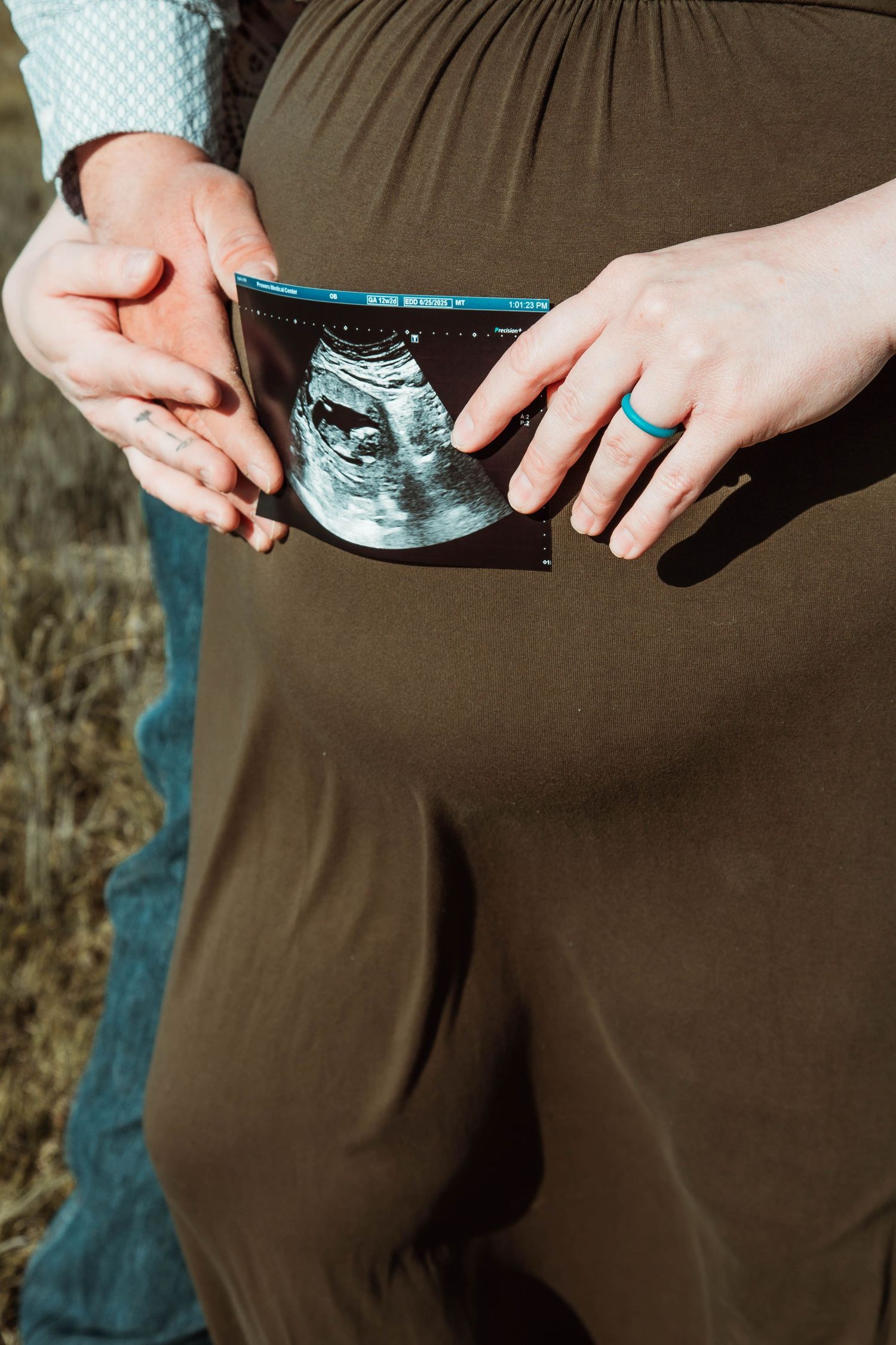 A pregnant woman in brown pants holds an ultrasound photo, with a teal ring visible on their hand.