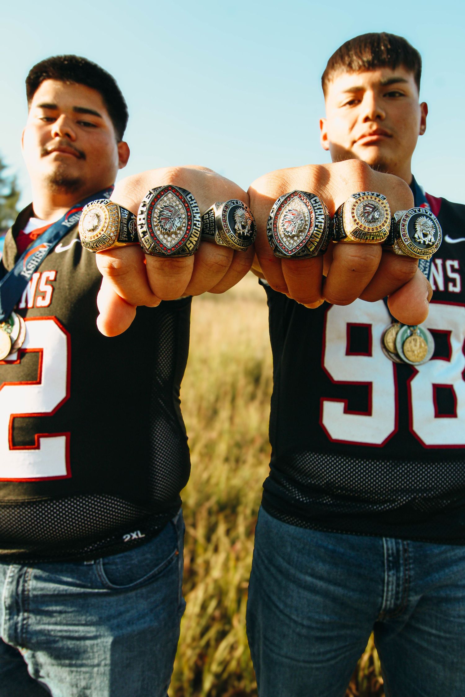 Five championship rings displayed in a row on hands against an outdoor background at sunset.