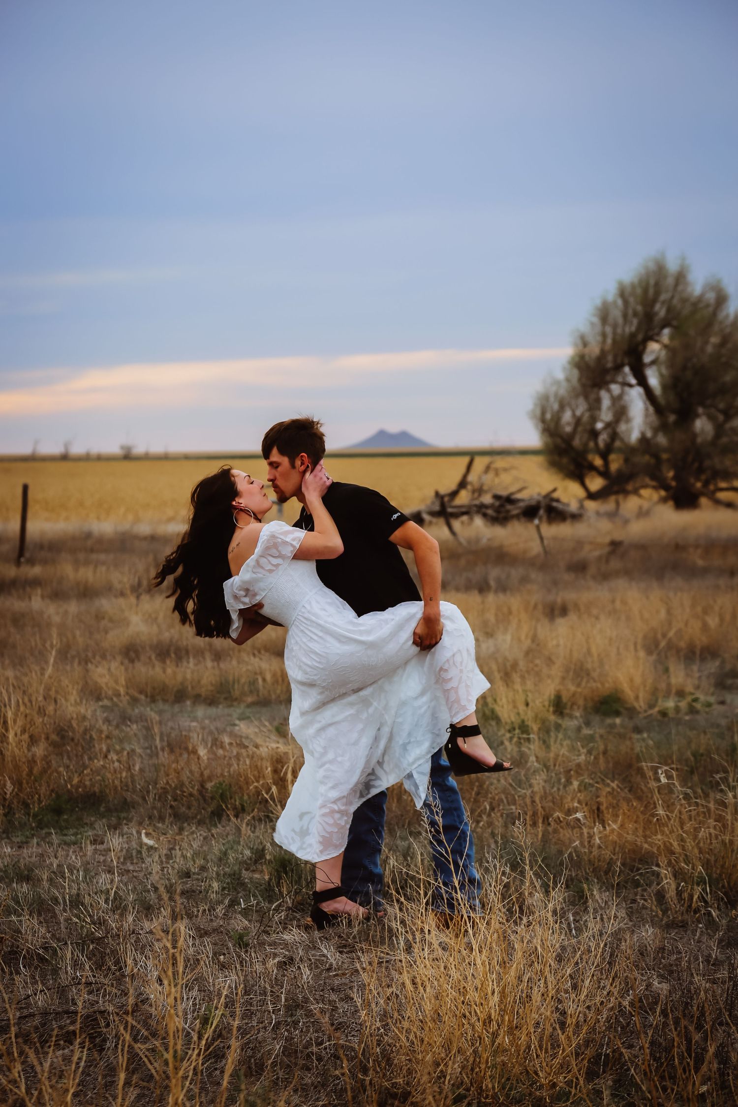 A romantic couple embraces in a golden field at sunset with mountains in the distance.