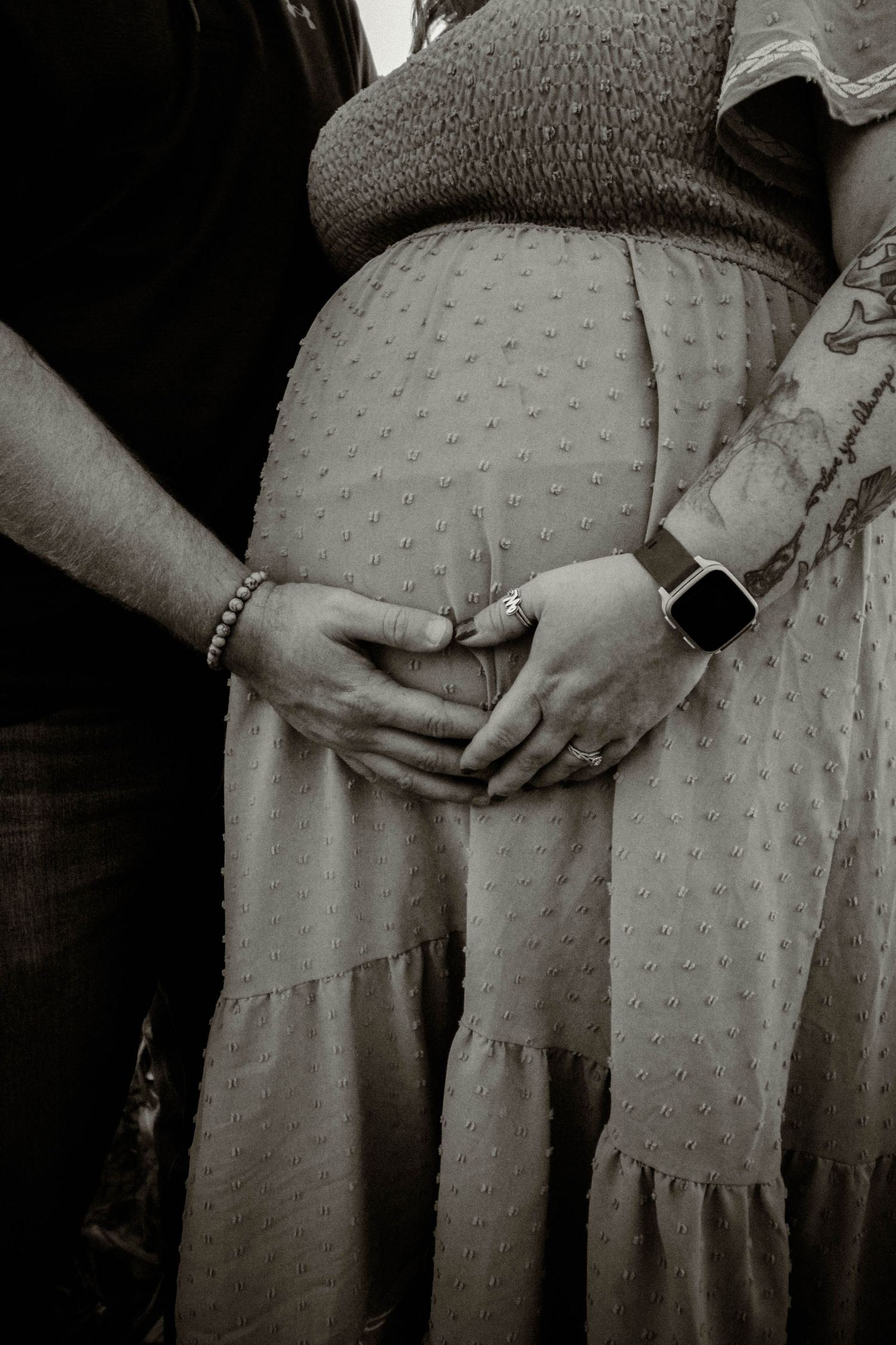 A black and white photo of hands forming a heart shape around a pregnant belly in a dotted dress.