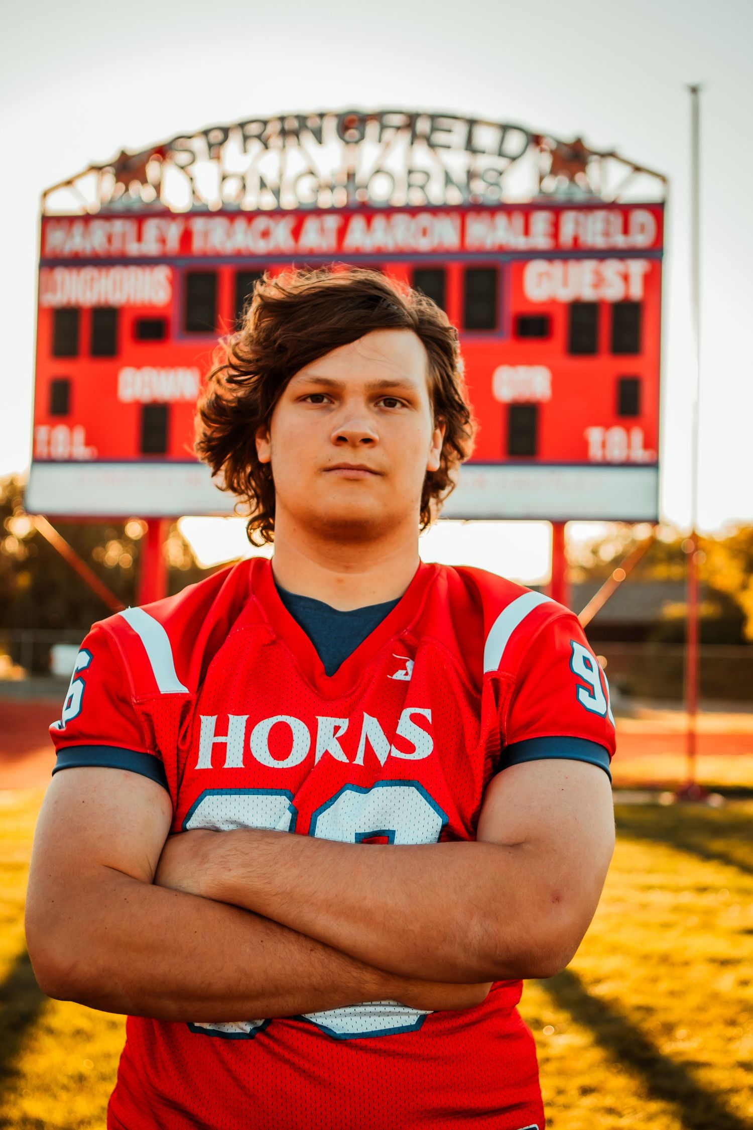 High school football player in red jersey poses at sunset on athletic field with scoreboard in background.