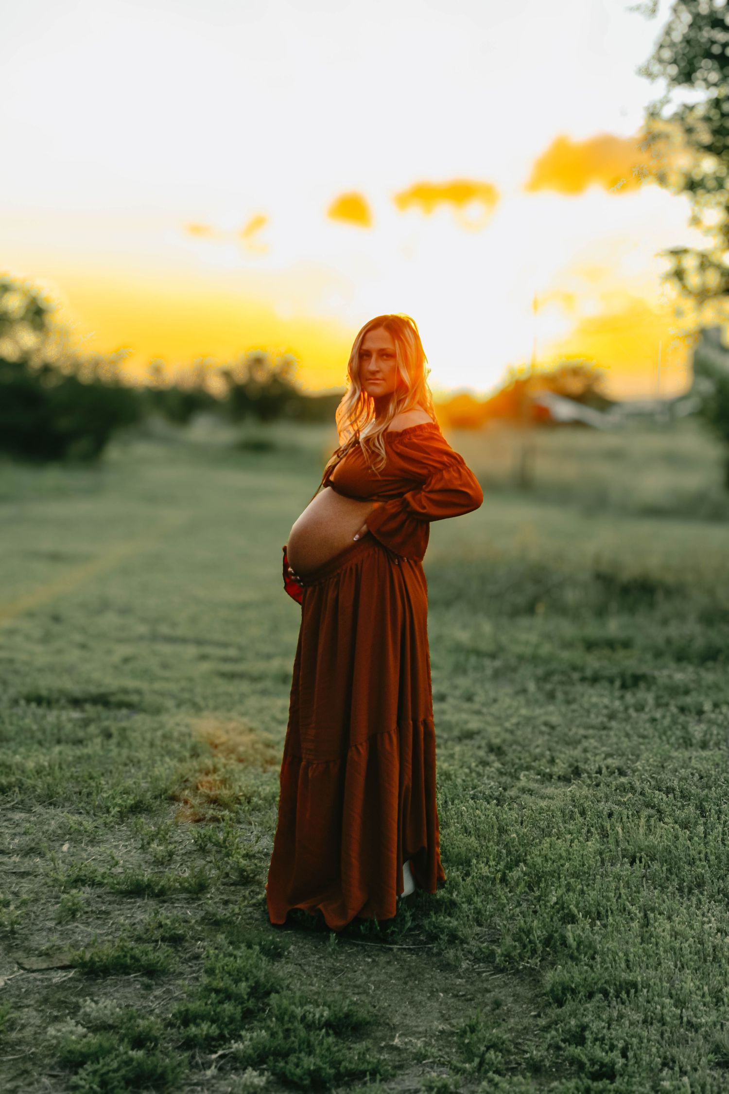 A silhouette of a pregnant woman in a long dress stands in a field at sunset.