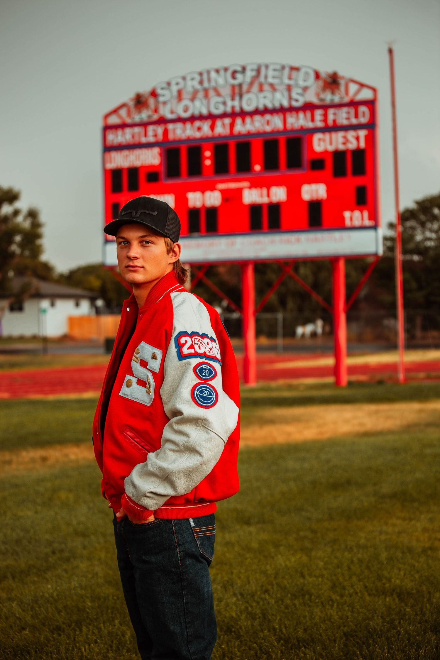 High school athlete poses in red and white varsity jacket near football stadium bleachers at sunset.