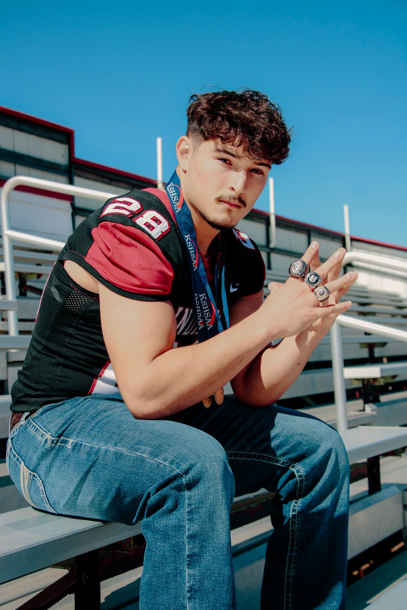 Person in jeans and dark shirt sits on bleachers while looking at mobile phone in outdoor stadium setting.