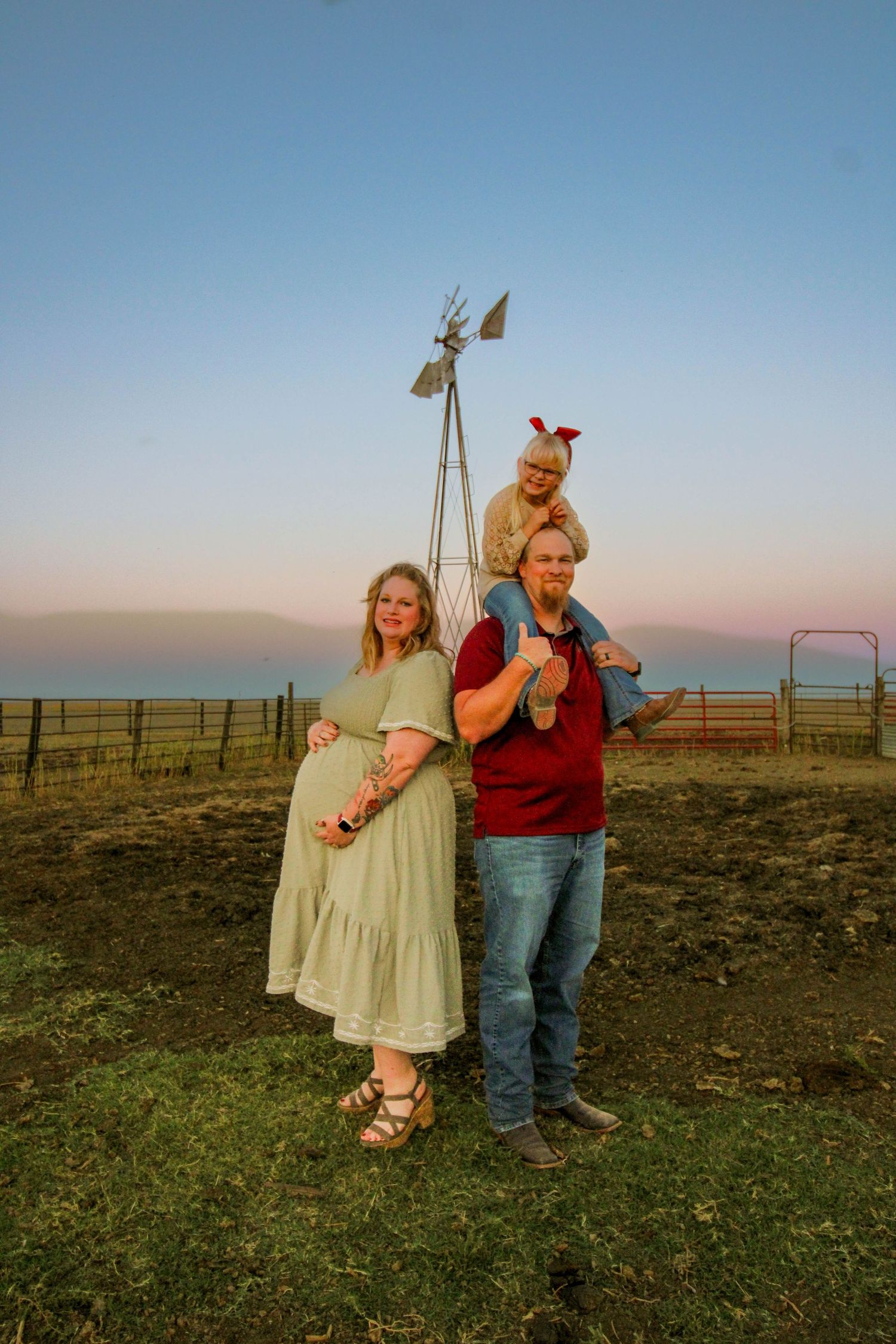A couple stands in front of a windmill on a farm during sunset, with the woman wearing a flowing dress.
