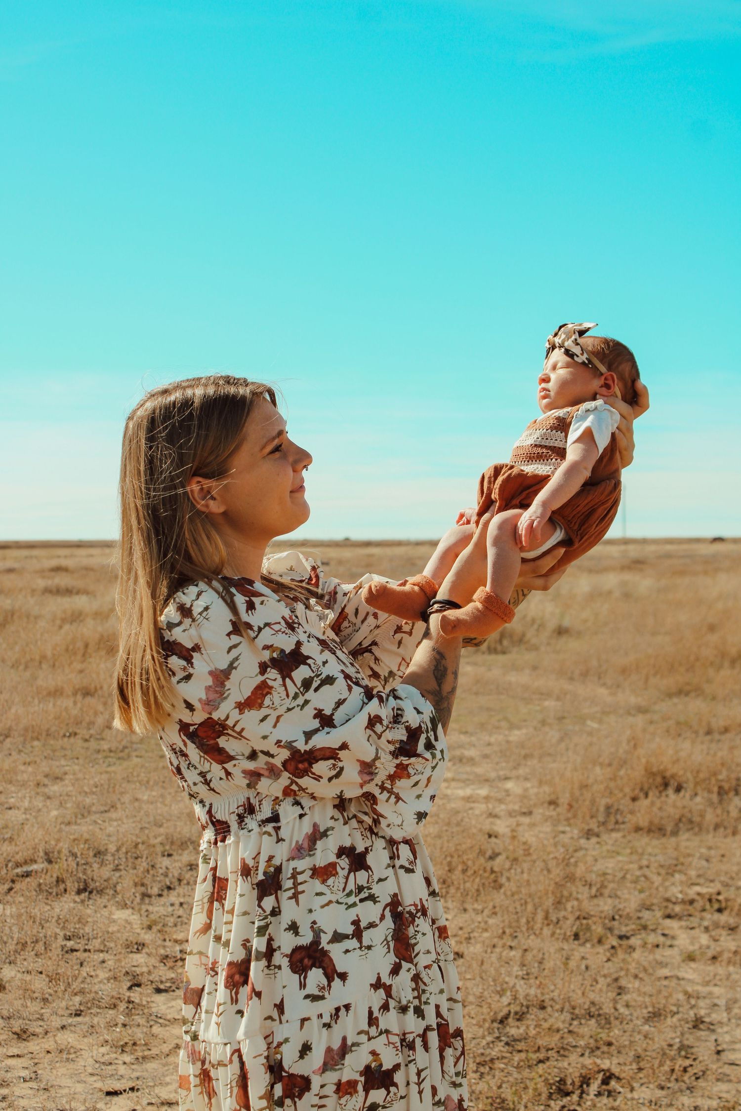 Woman in floral dress holds infant up in golden field against bright blue sky during sunset.