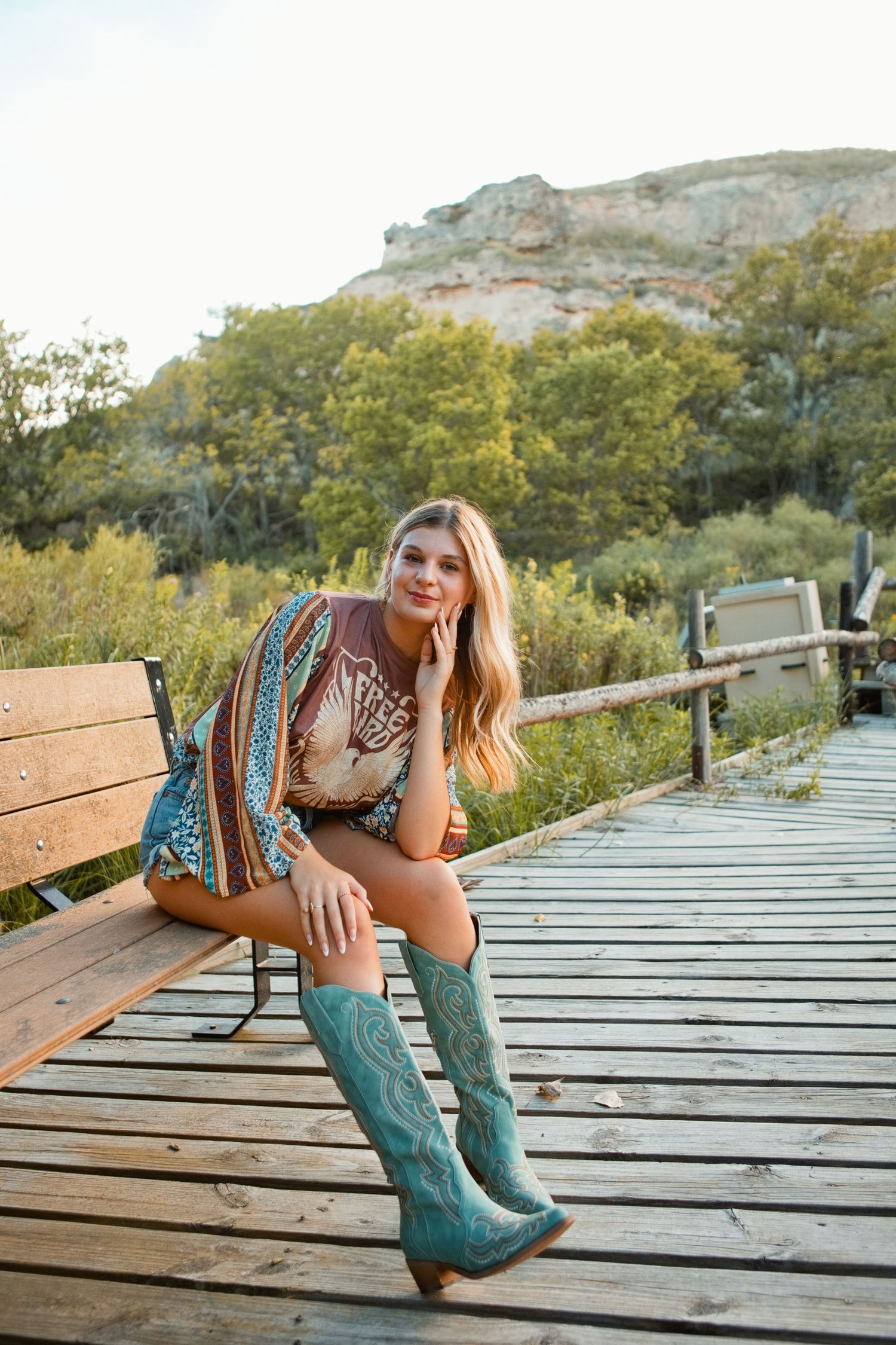 Young woman in teal cowboy boots and boho outfit sits on wooden boardwalk bench with rocky hills and trees behind her.