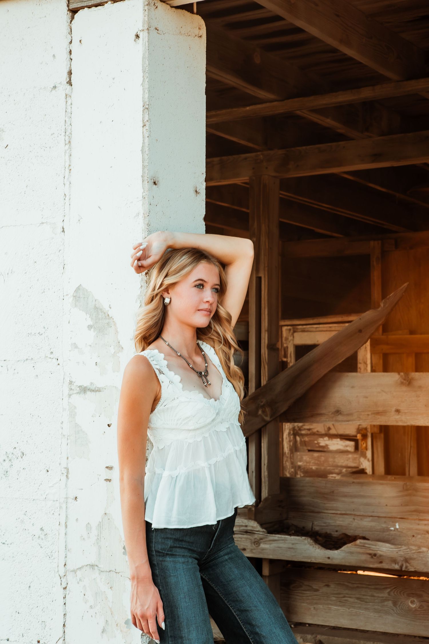 Young blonde woman in white lace top and jeans poses gracefully against rustic wooden barn structure.