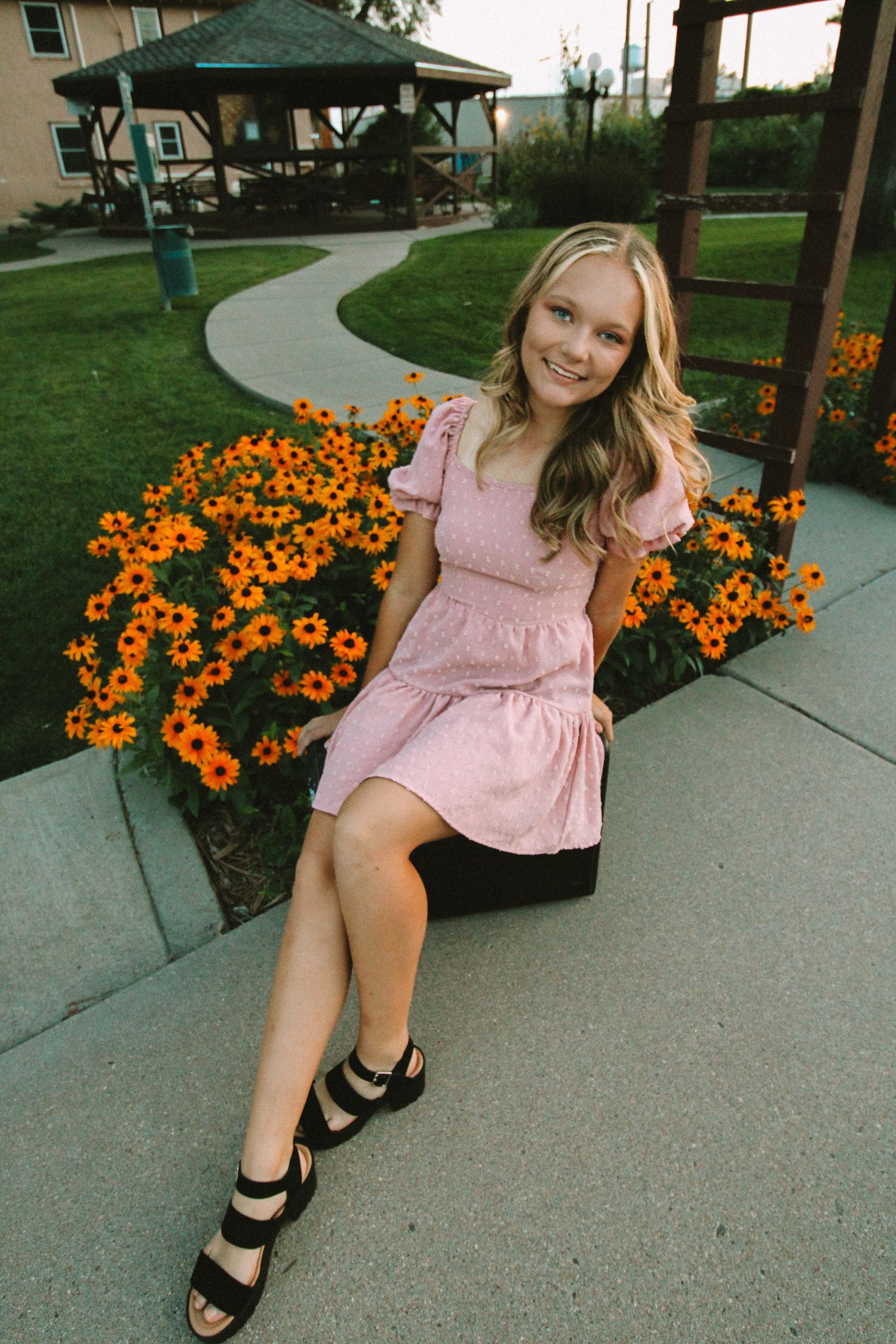 A person in a pink dress sits on a concrete path near vibrant orange flowers in a garden setting.