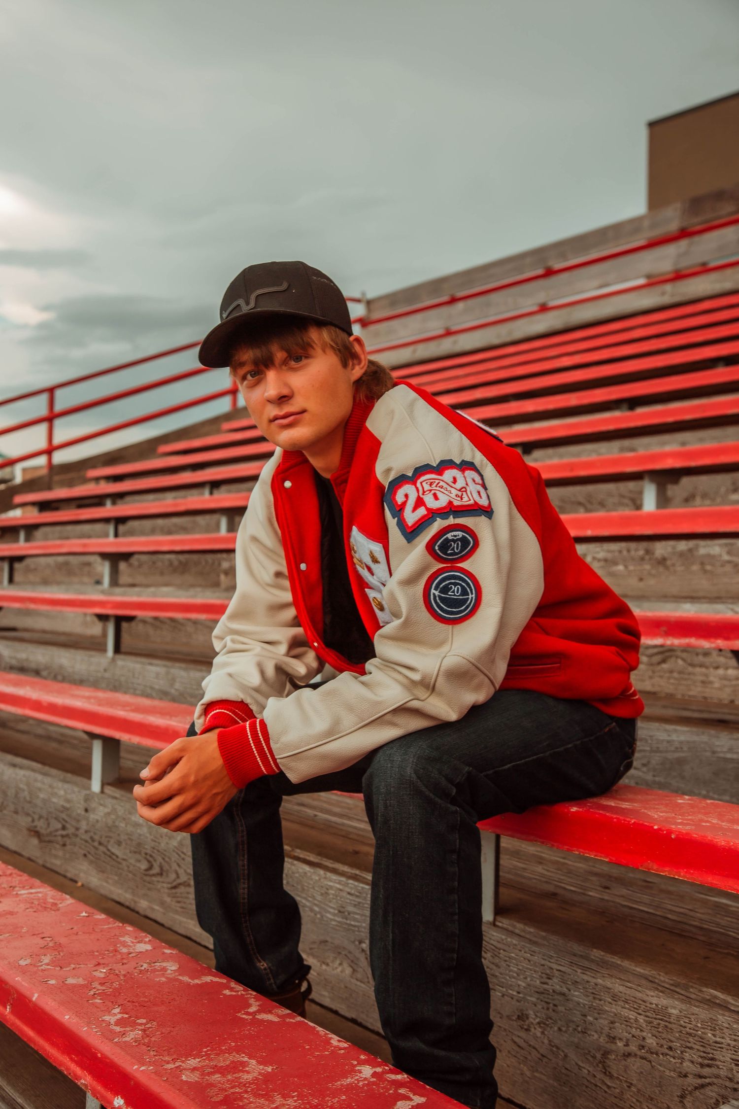 Person wearing red and white varsity jacket sits on red bleachers at a stadium during overcast day.