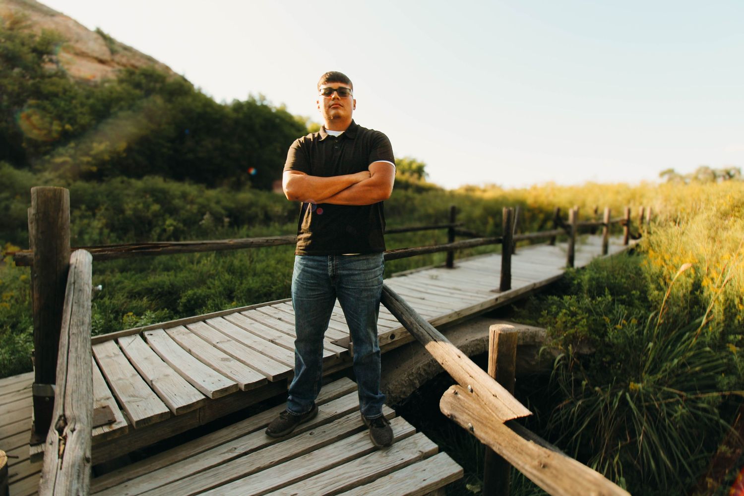 A person stands on a wooden boardwalk path through tall grasses at sunset in a nature preserve.