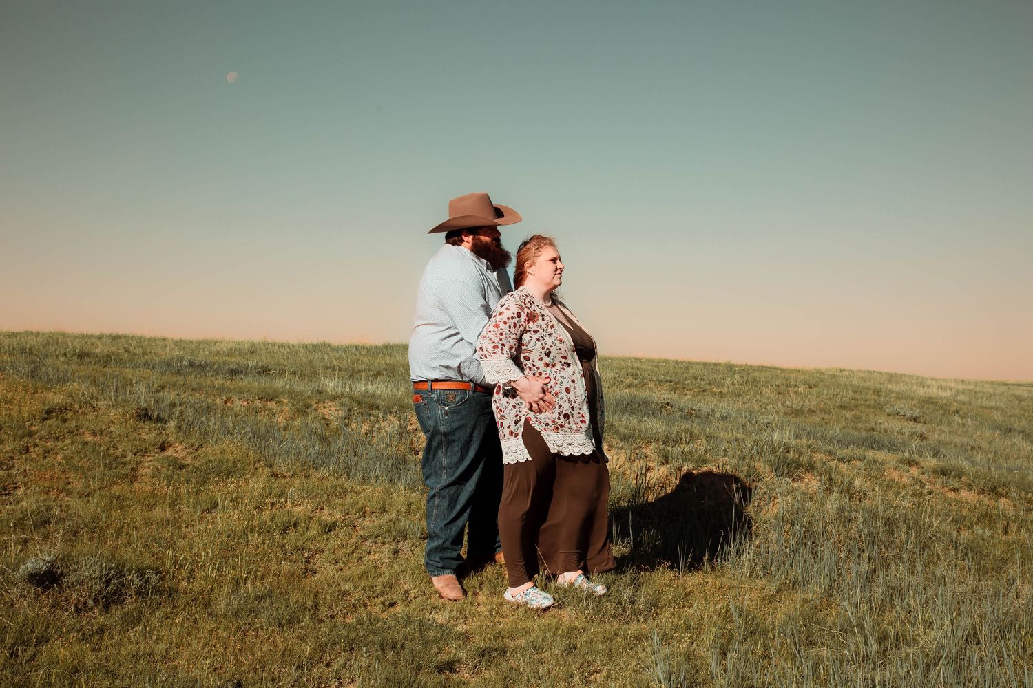 Two people stand together on rolling grassland hills under a pastel sunset sky.