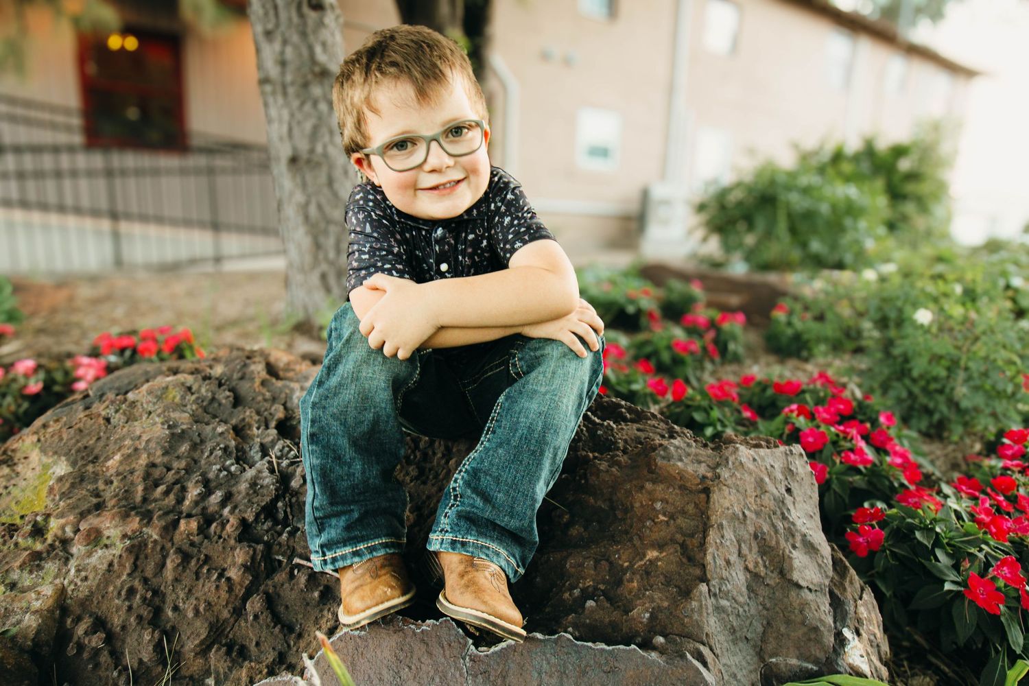 Young person in glasses sits confidently on a large rock surrounded by vibrant red flowers in a garden setting.