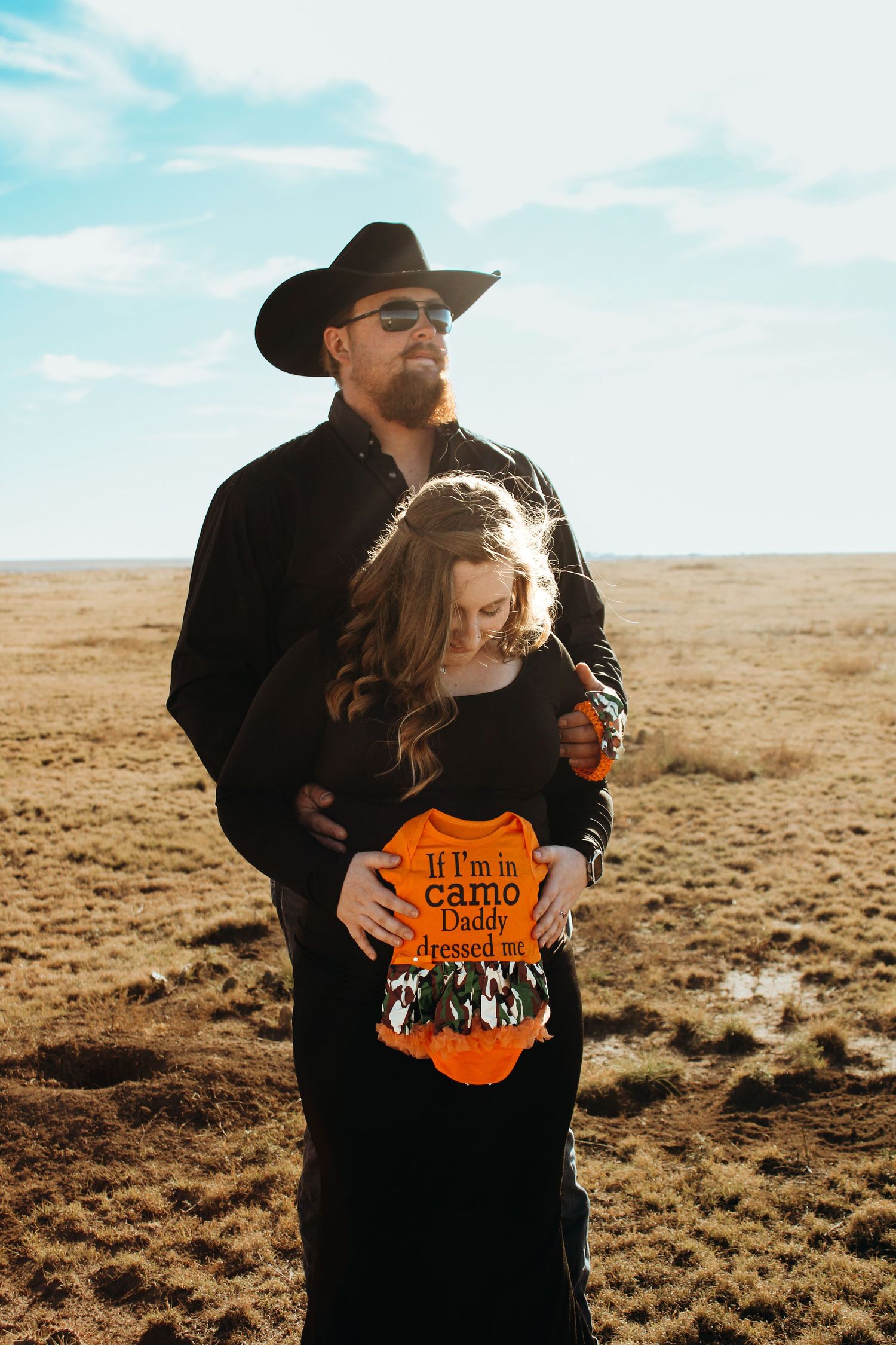 Two people stand together wearing black clothing in an open desert landscape against a blue sky with white clouds.